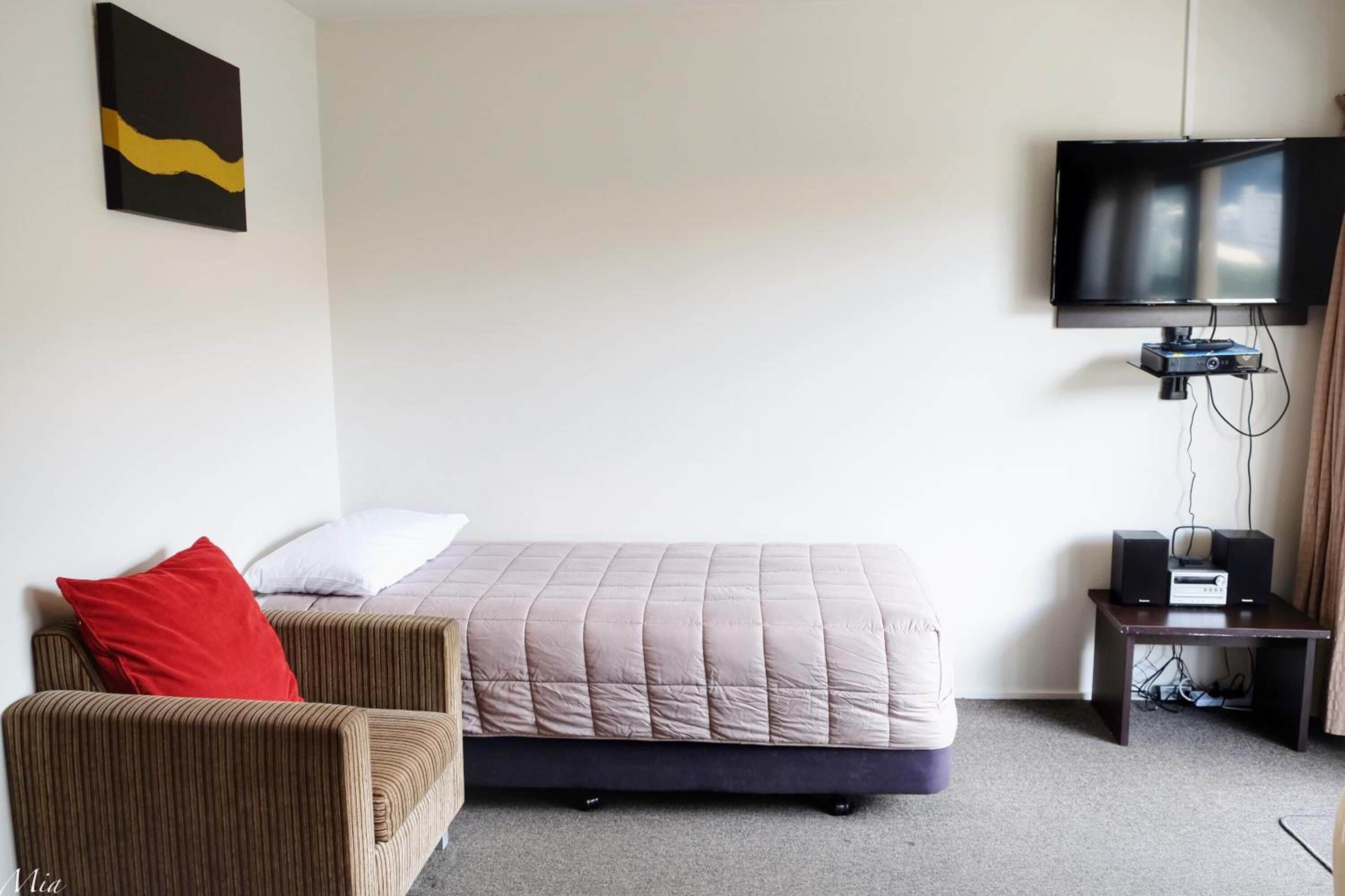 Una habitación de hotel con una cama, una silla, un televisor y una pequeña mesa con altavoces.