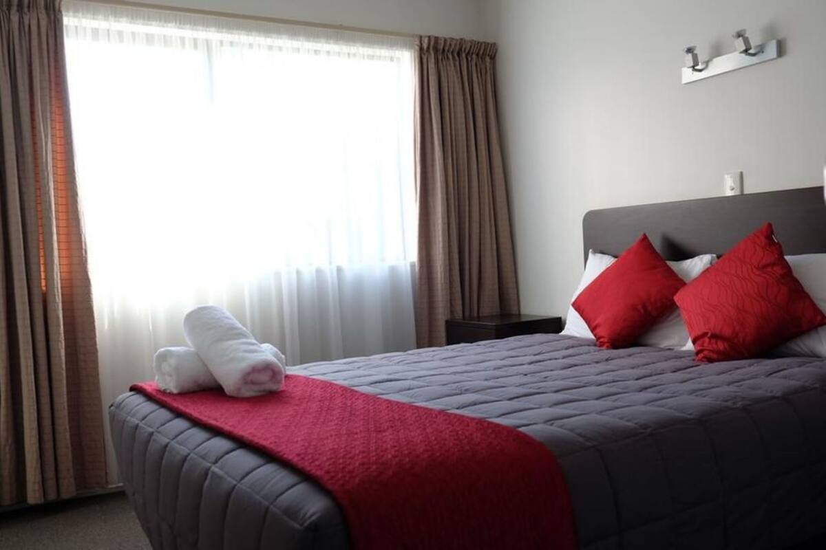 A hotel room with a bed, red pillows, a red blanket, and a window with curtains.