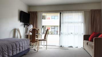 Una habitación de hotel con una cama, un escritorio con una silla, un sofá con cojines rojos, un televisor montado en la pared y un gran ventanal con cortinas.
