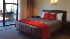 A hotel room with a large bed, a nightstand, a wall-mounted lamp, and a sliding glass door leading to an outdoor area. - Marksman Motor Inn (Wellington)