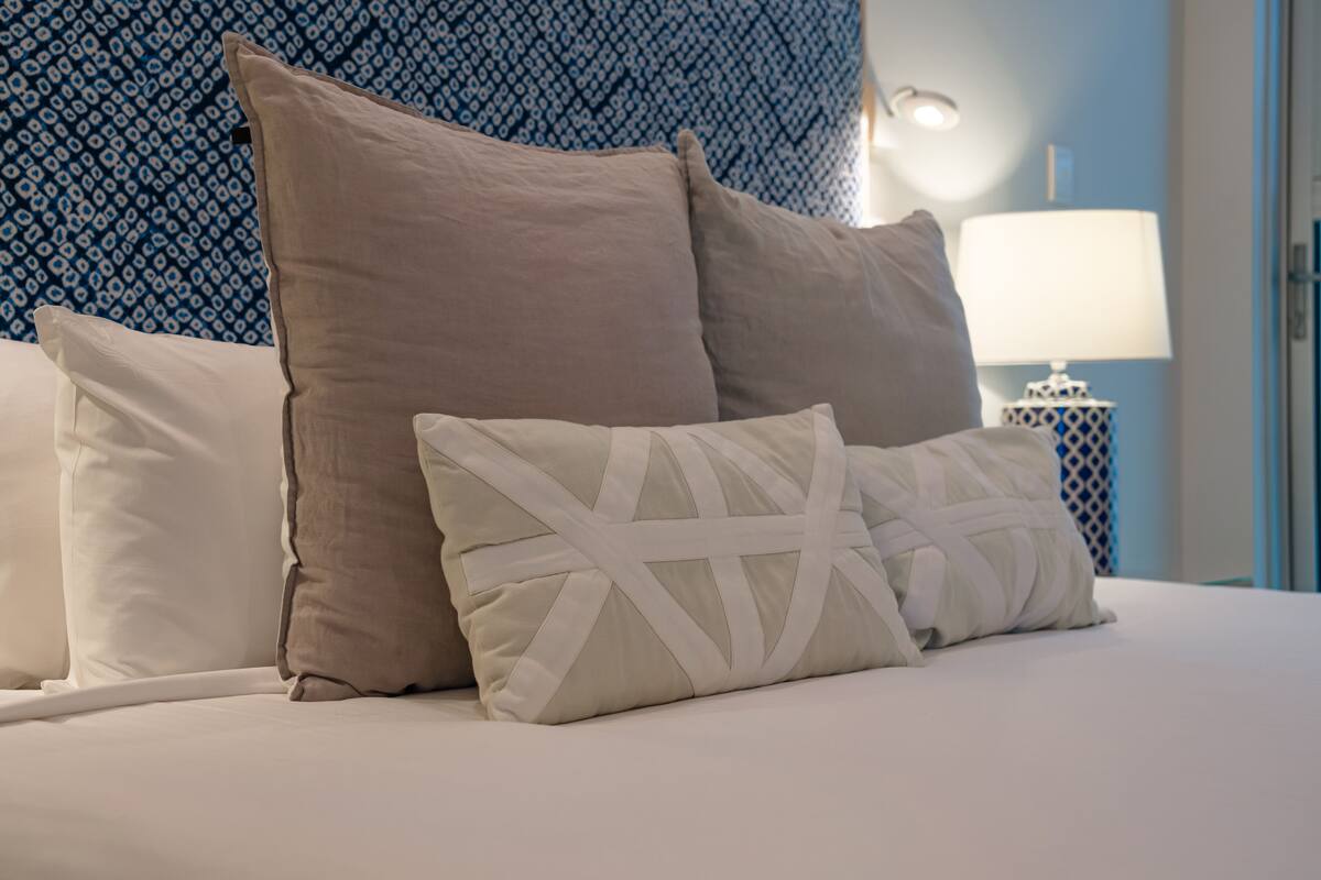 A bed with beige and white pillows, a patterned blue wall, and a bedside table with a lamp.