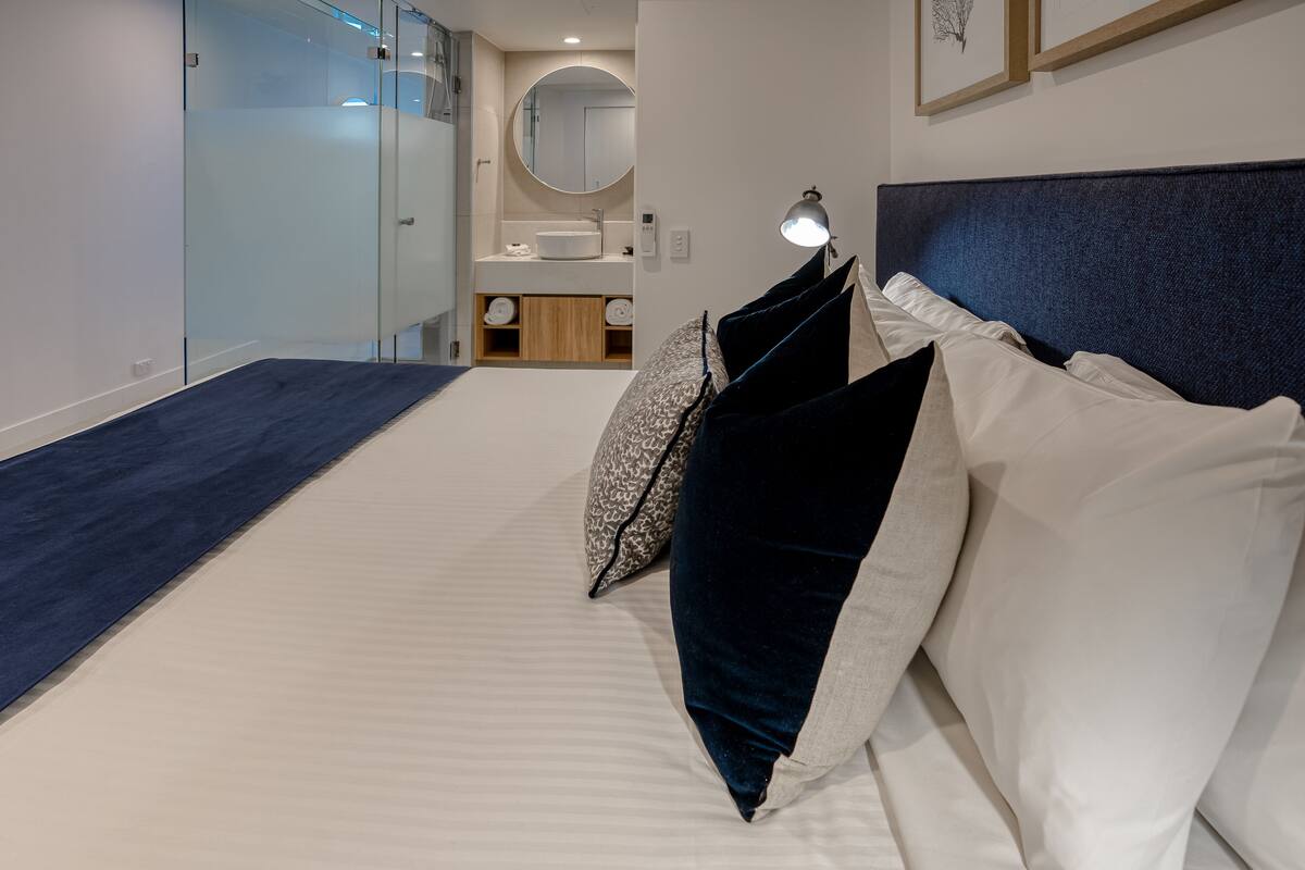 A modern bedroom with a large bed, a bedside table with a lamp, a mirror, and a bathroom visible through a glass door.