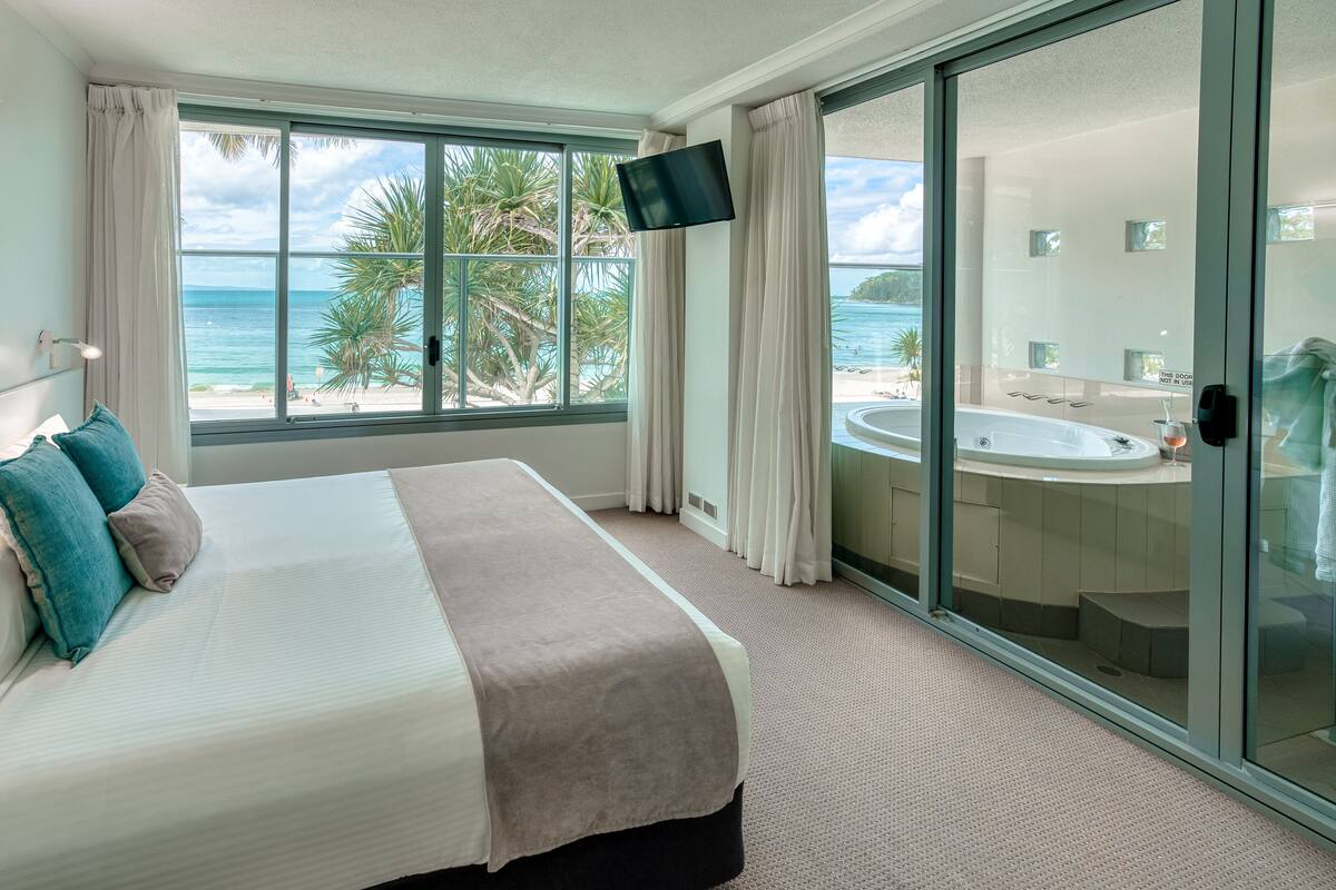 A hotel room with a bed, a TV, a large window with a view of the beach and ocean, and a jacuzzi.