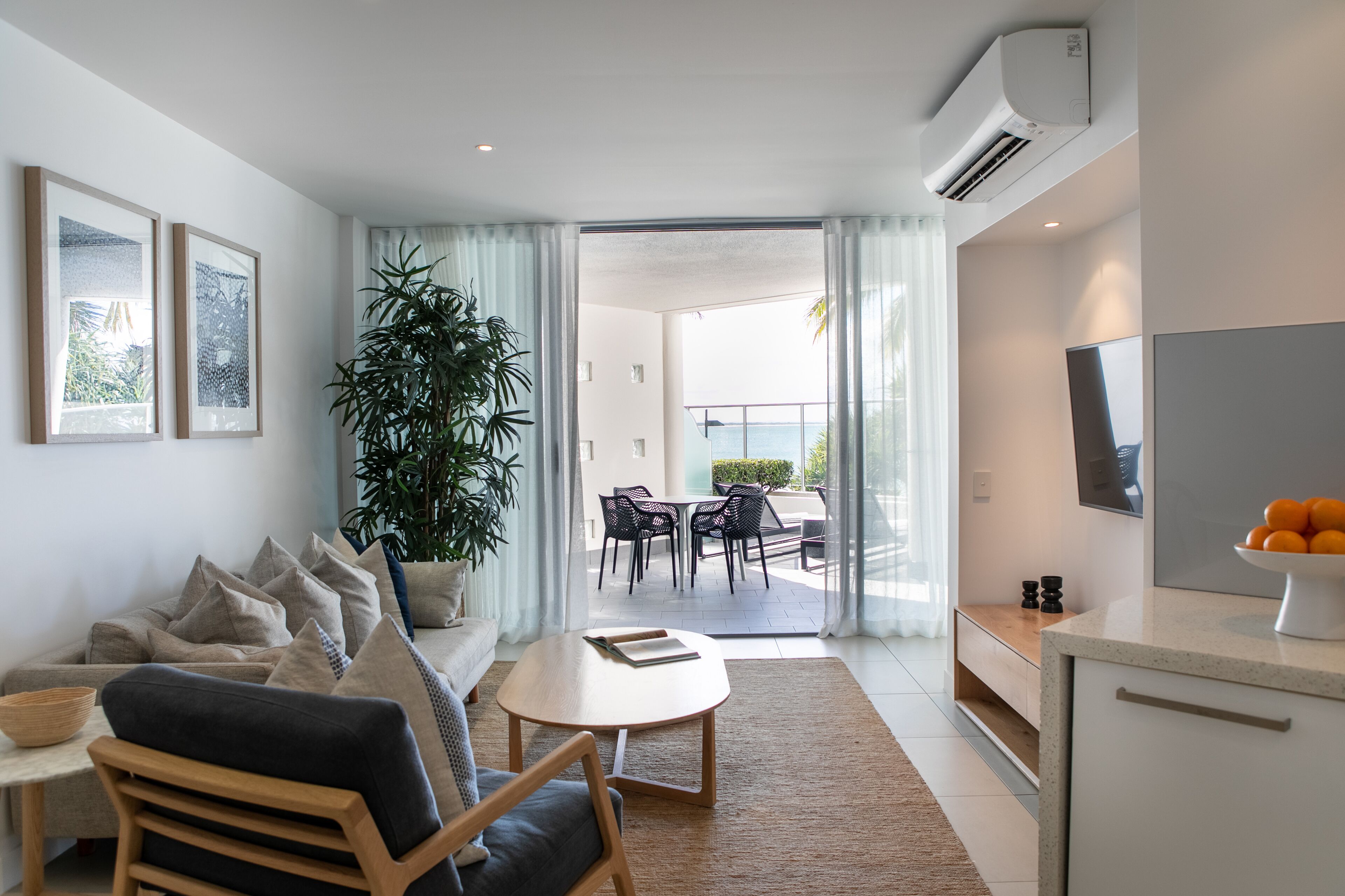 Een moderne woonkamer met een bank, salontafel en een grote potplant. Er is een eethoek met stoelen en een tafel, en een keuken met een televisie en een schaal fruit.