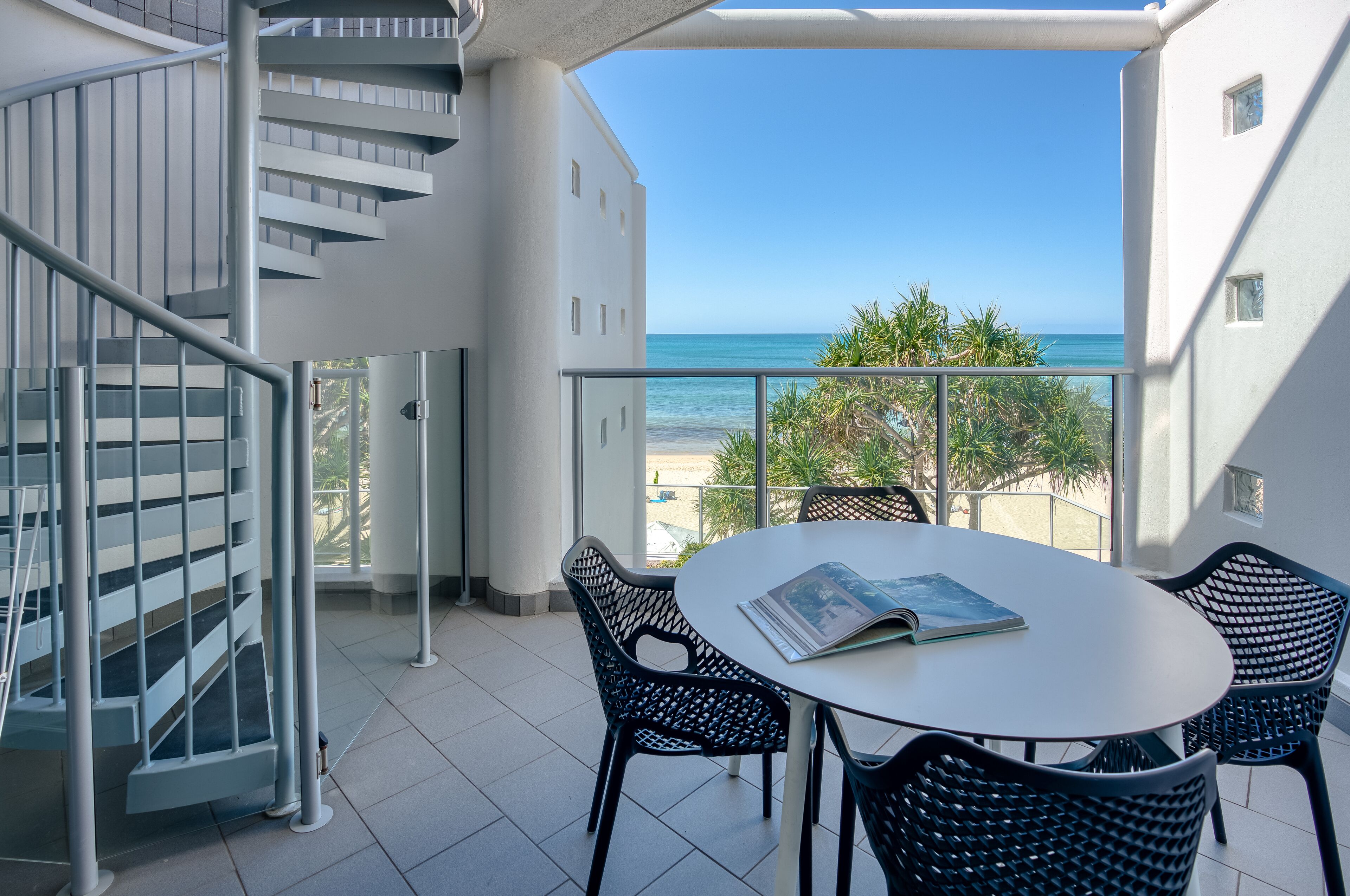 Un balcon agrémenté d’un escalier en colimaçon, une table ronde avec des chaises et une vue sur la plage.