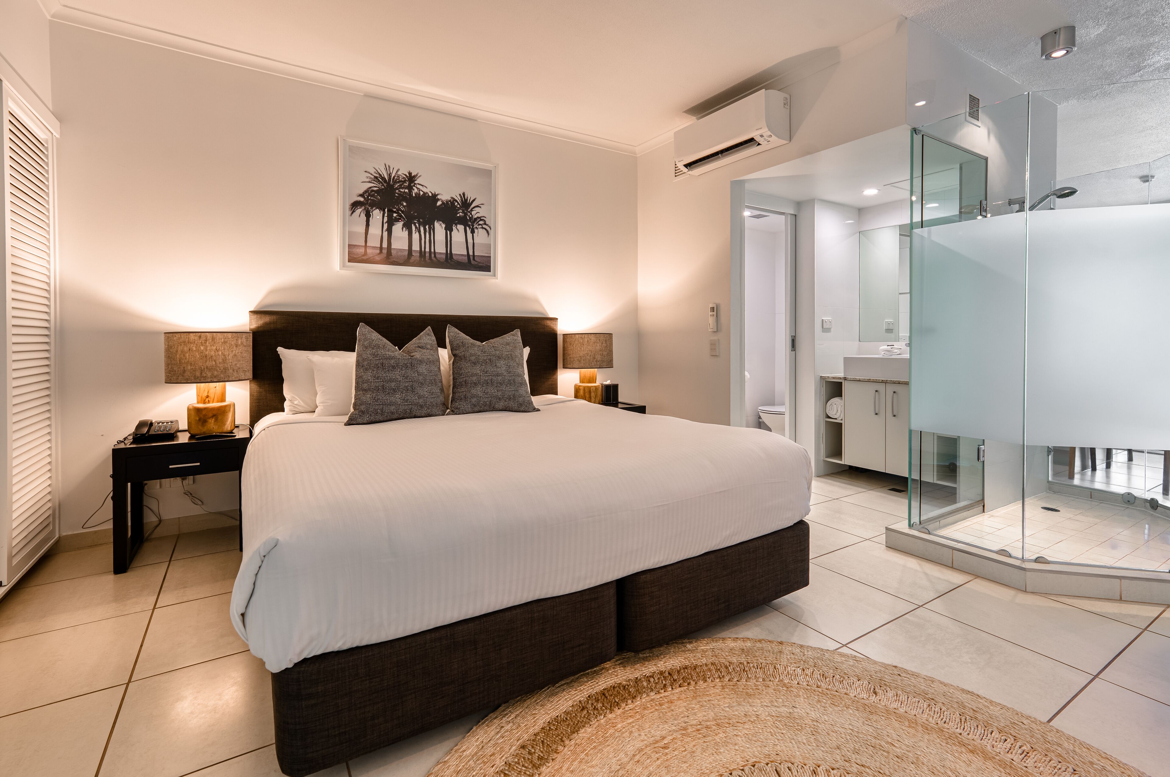 A modern bedroom with a large bed, bedside lamps, a nightstand, a wall-mounted air conditioner, and a glass-enclosed shower area.
