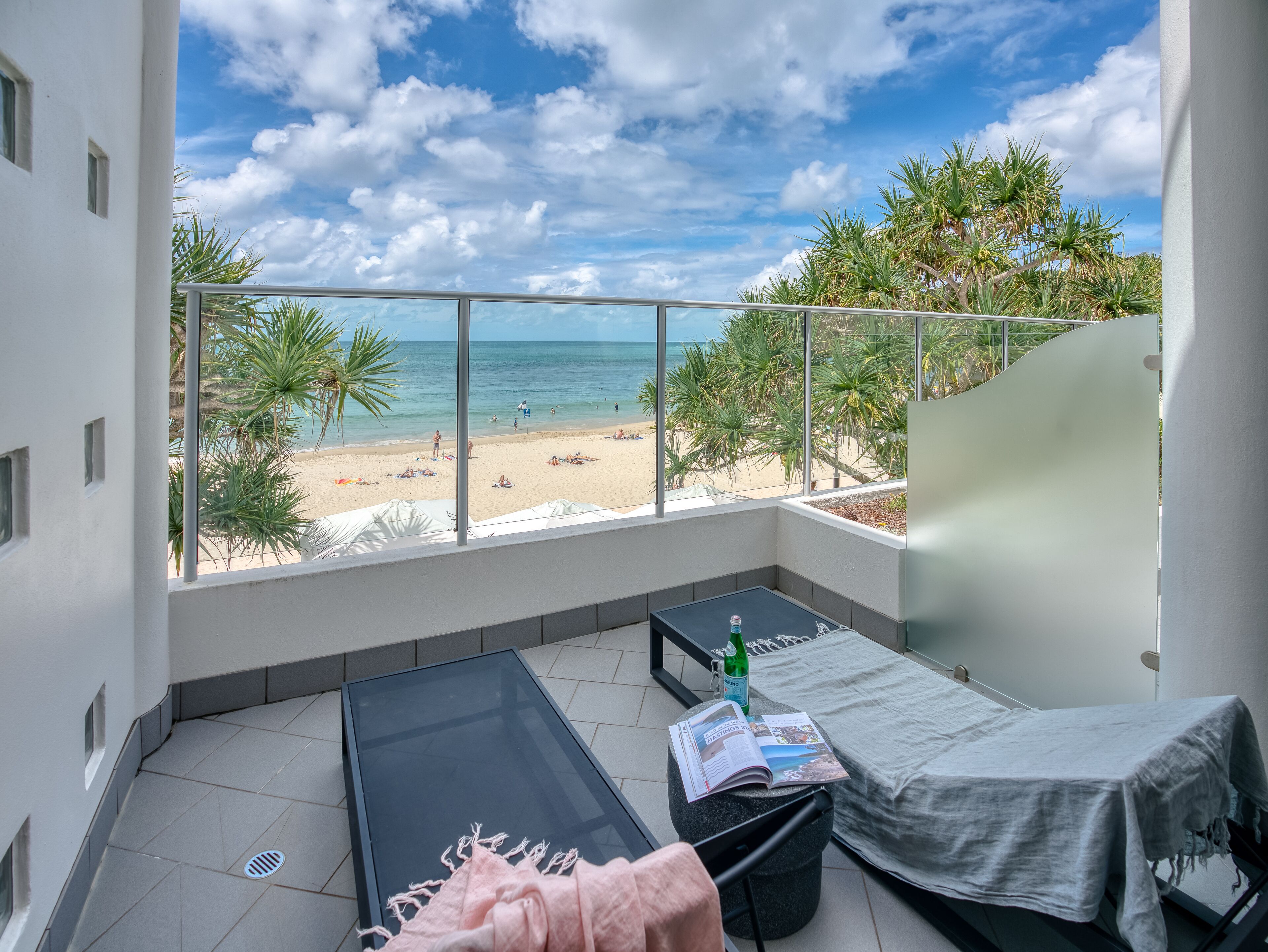 A balcony with a view of a beach, a bed, a table with a magazine, and a bottle of water.