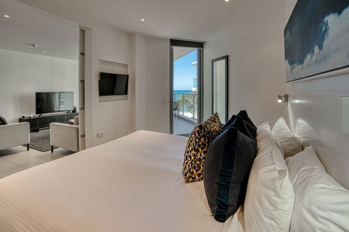 A modern hotel room with a large bed, a TV, a seating area, and a balcony with a view of the sea.