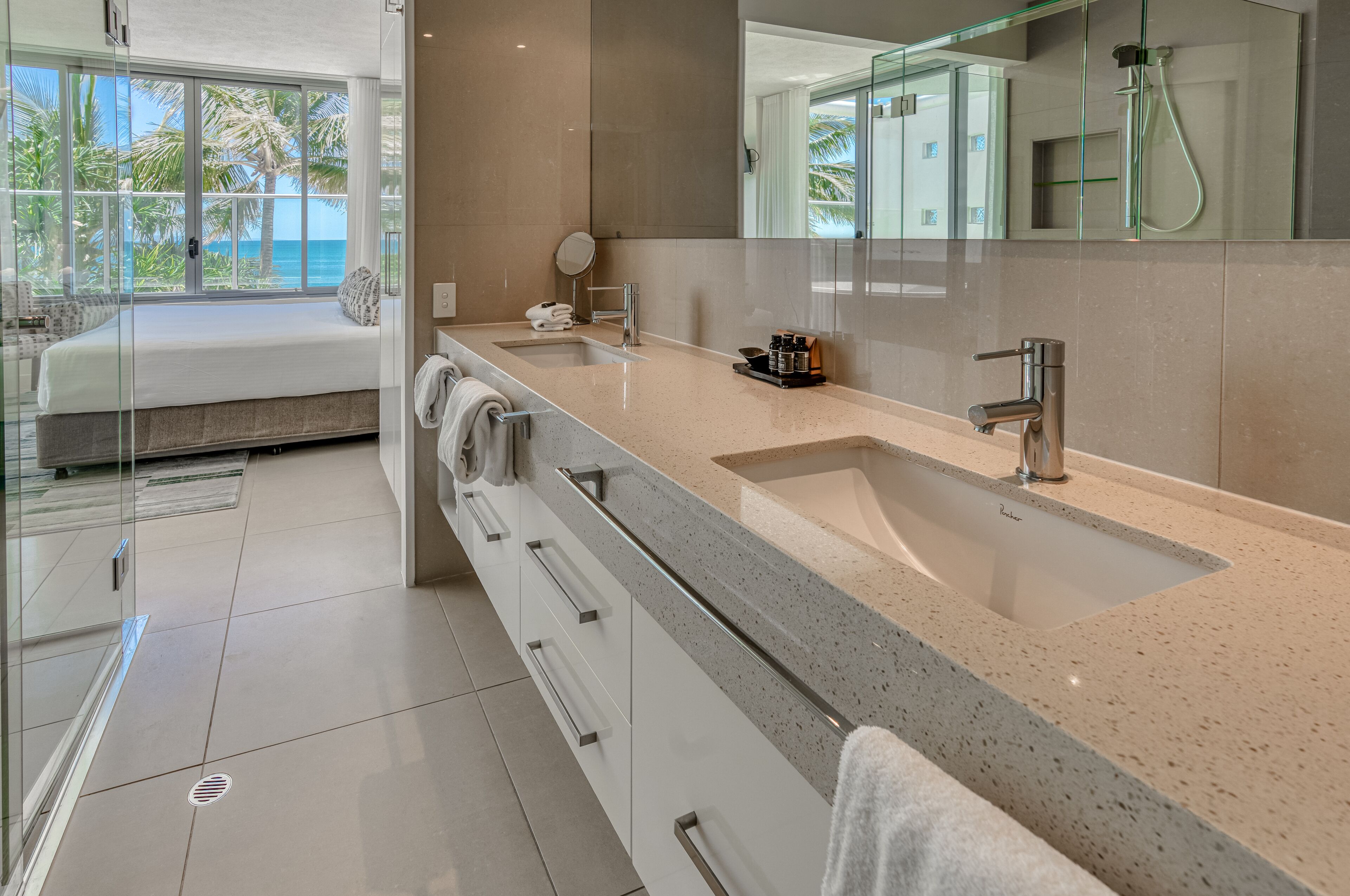 Ein modernes Badezimmer mit einem großen Spiegel, zwei Waschbecken und einer Dusche. Ein Bett mit Blick auf Palmen und das Meer.