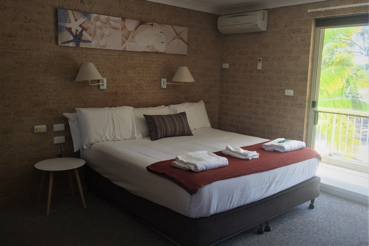 A hotel room with a brick wall, a bed with white linens, two wall-mounted lamps, an air conditioner, and a window with a view of palm trees.