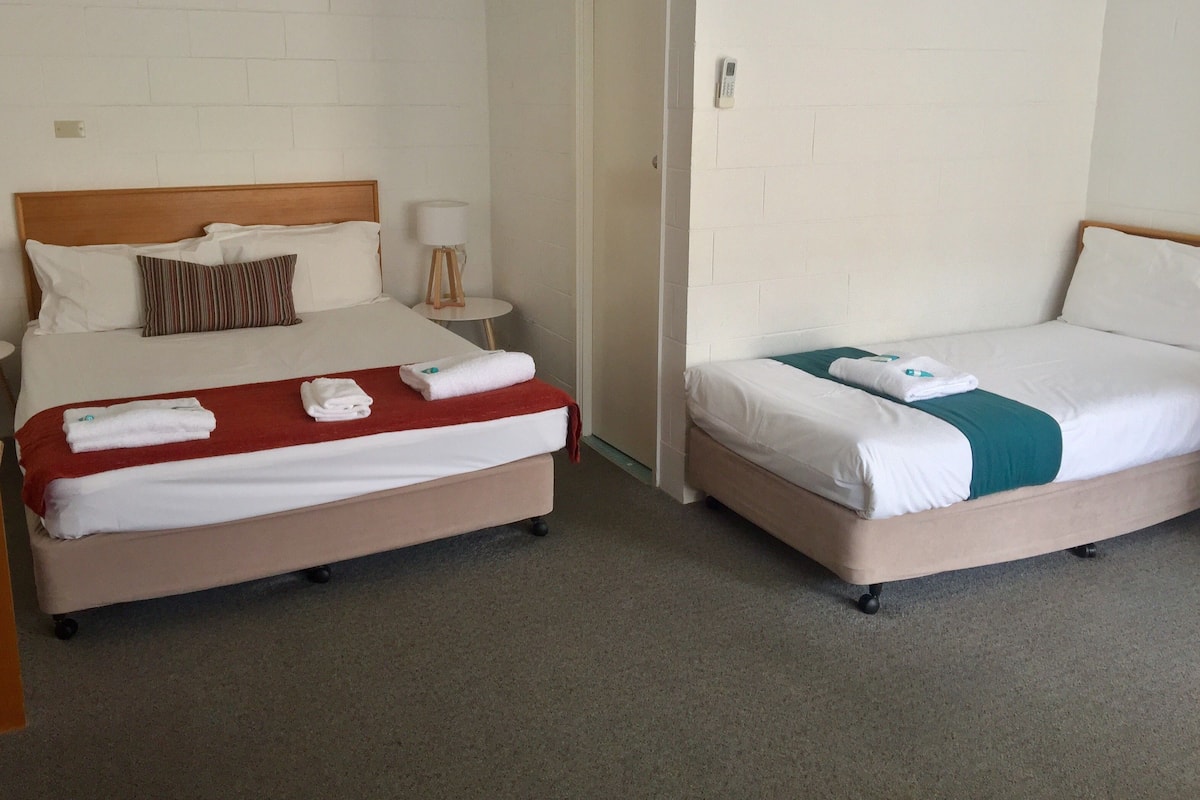A hotel room with two beds, each with white linens and a red and green blanket. There is a small bedside table with a lamp and a wall-mounted phone.