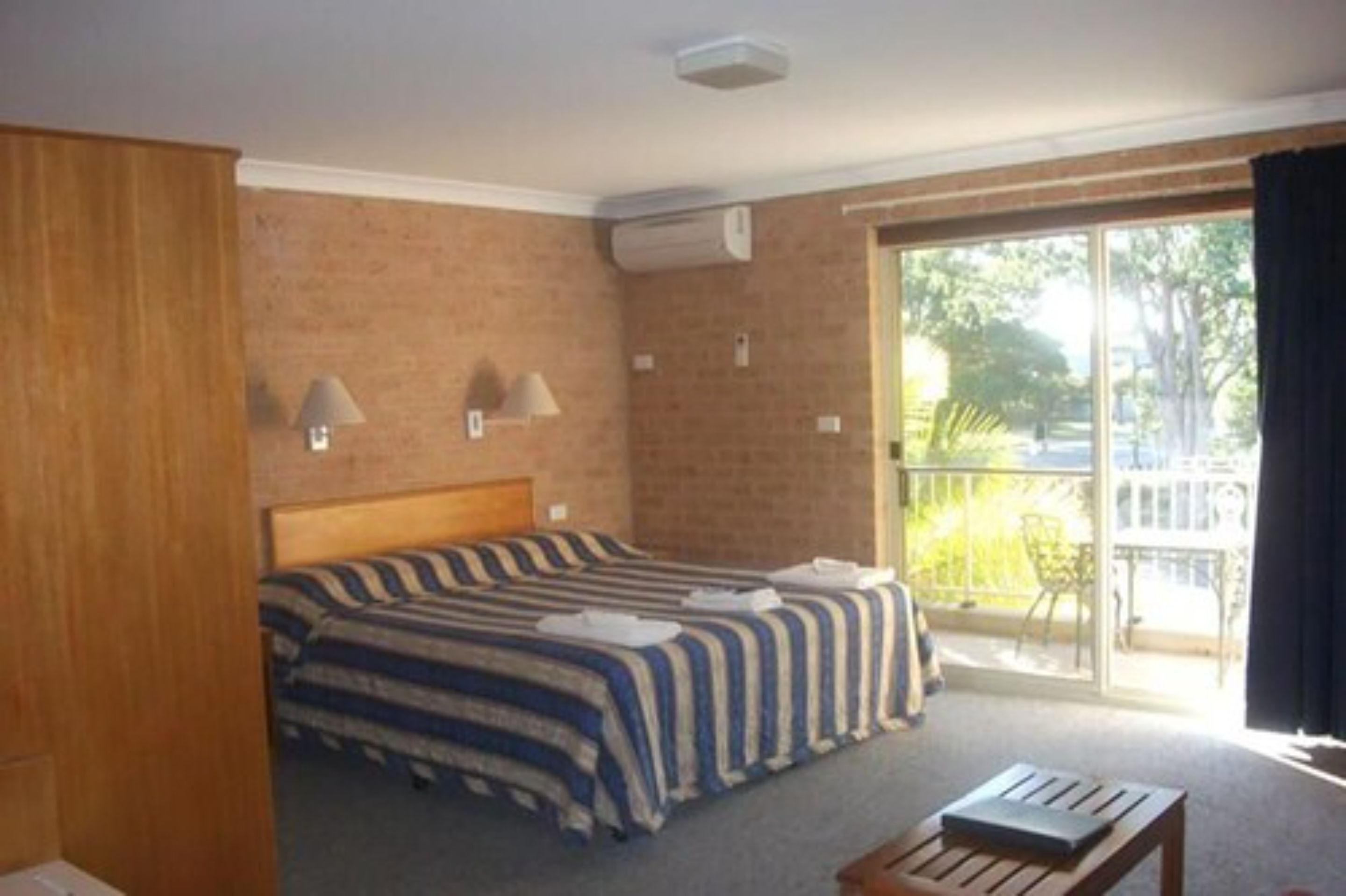 A hotel room with a large bed, a wooden headboard, a small table, and a balcony with a view of greenery.