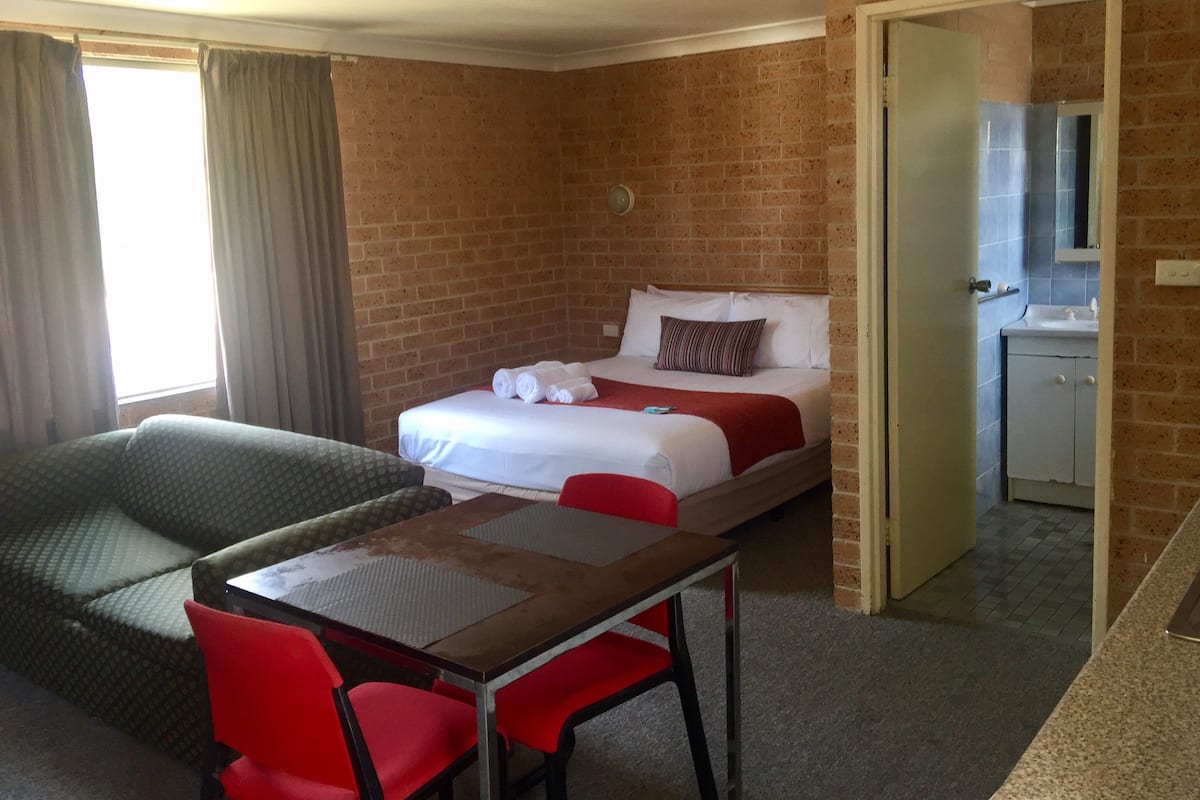 A hotel room with a bed, a small table with two chairs, a sofa, and a bathroom visible through an open door.