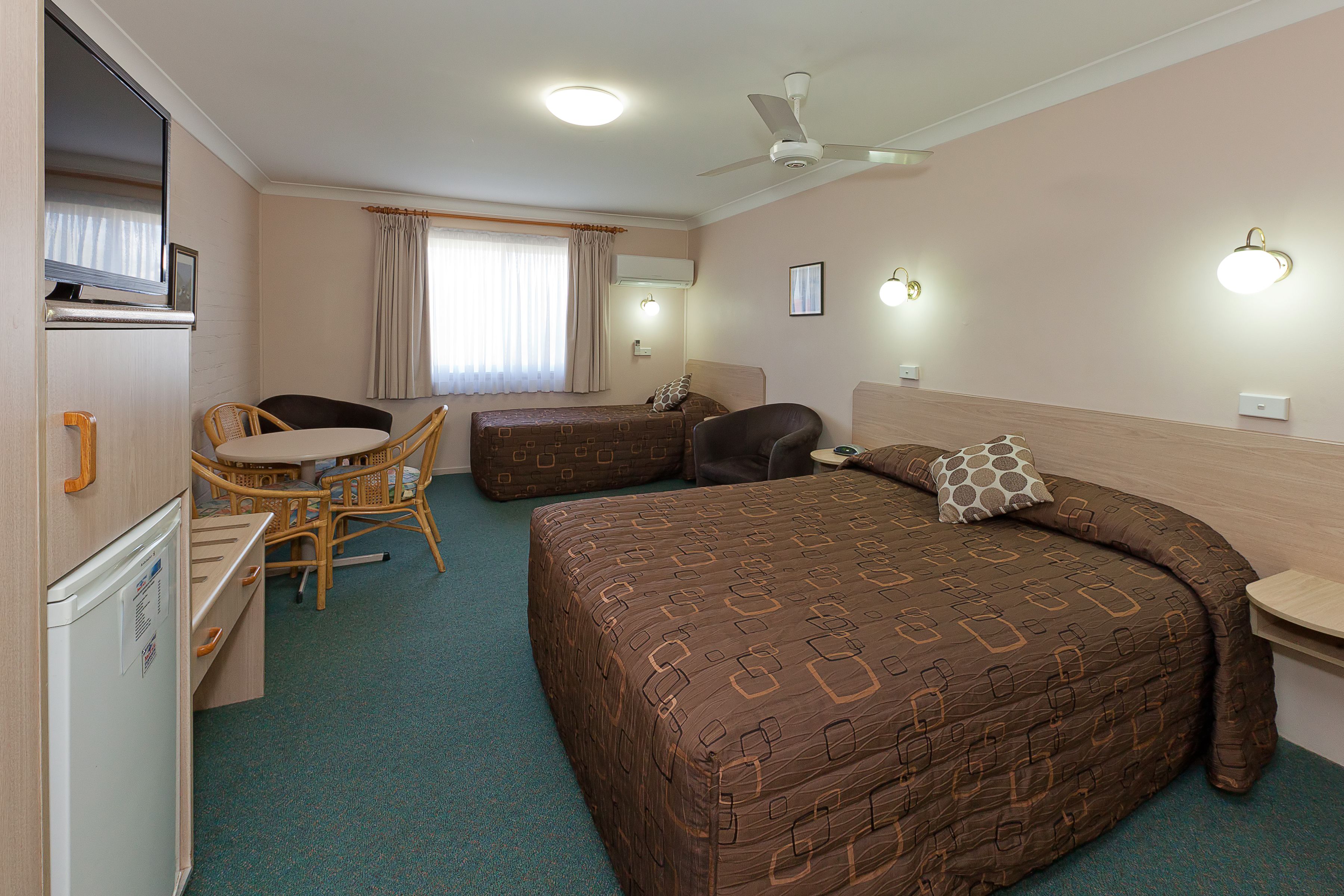 A hotel room with a bed, a desk, a chair, a small table, a ceiling fan, and a window with curtains.