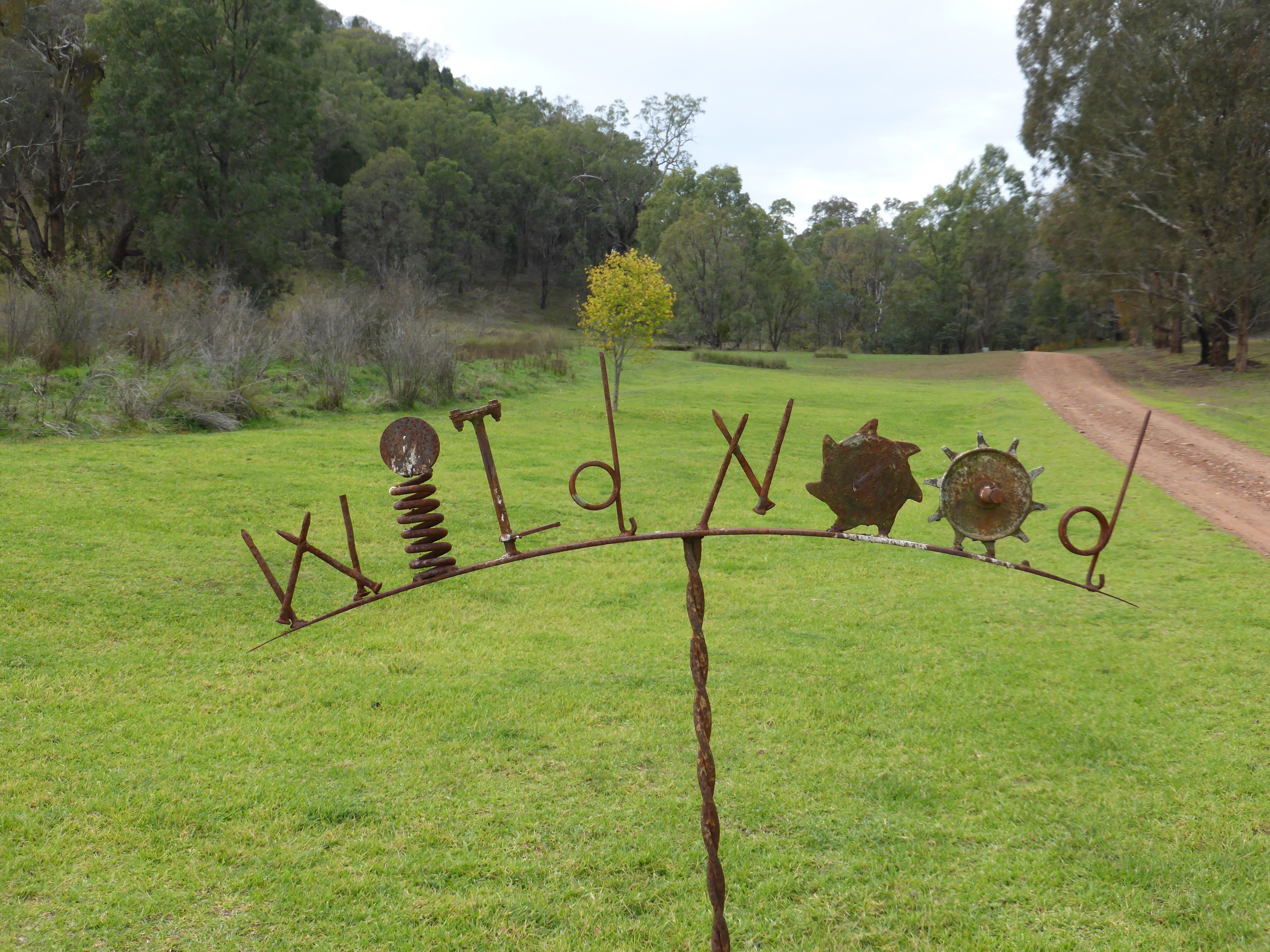 A rustic metal sculpture in a grassy field with a dirt path in the background.