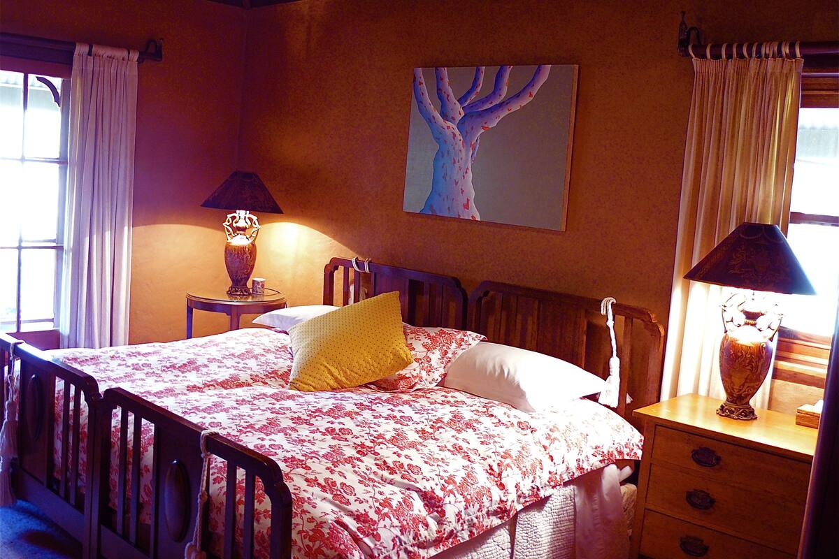 Een slaapkamer met een bed, twee nachtkastjes, een stoel, een schilderij aan de muur en een raam met gordijnen.