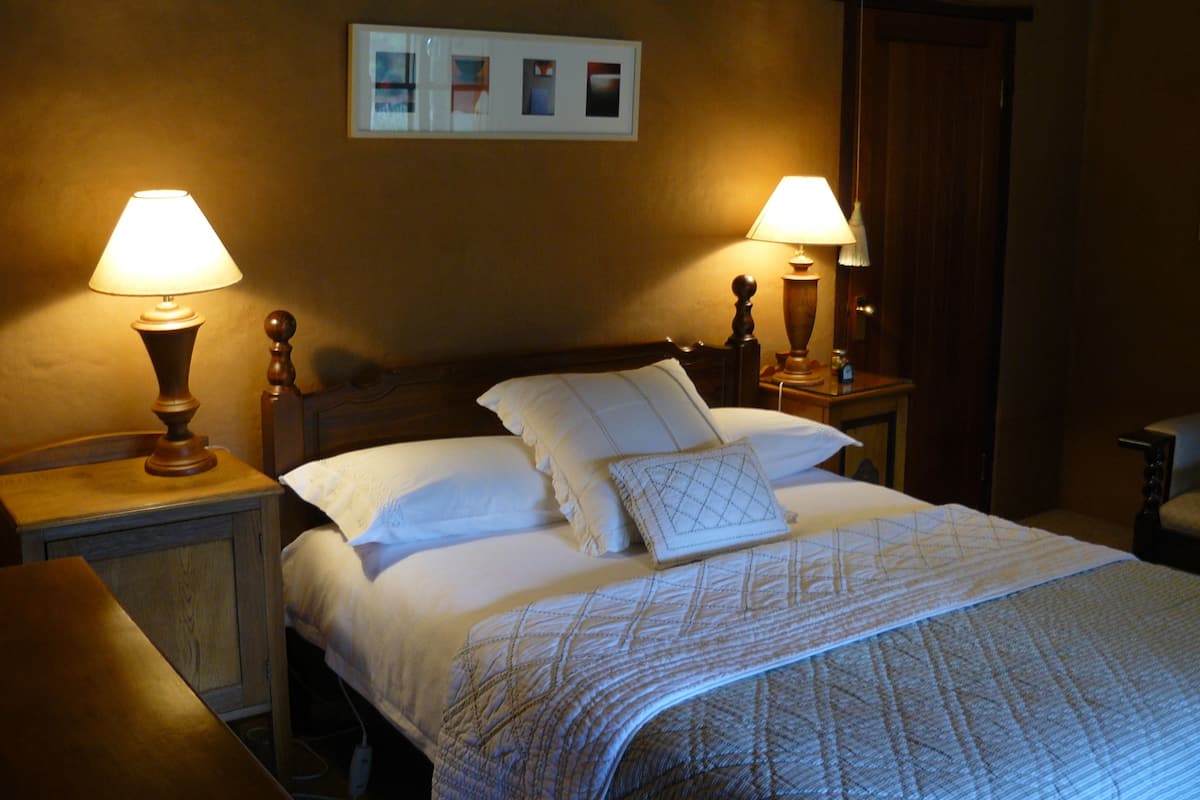 Een net opgemaakt bed met witte lakens en een gestreept deken, twee bedlampen en een ingelijst schilderij aan de muur erboven.