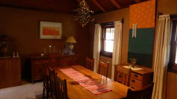 Una mesa de comedor de madera con sillas, una araña, una ventana con cortinas y un radiador fijado a la pared.