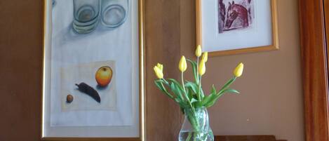 Une œuvre d’art encadrée représentant une nature morte avec des bouteilles et des fruits, une petite image encadrée, et un vase contenant des tulipes jaunes, le tout posé sur un meuble en bois.