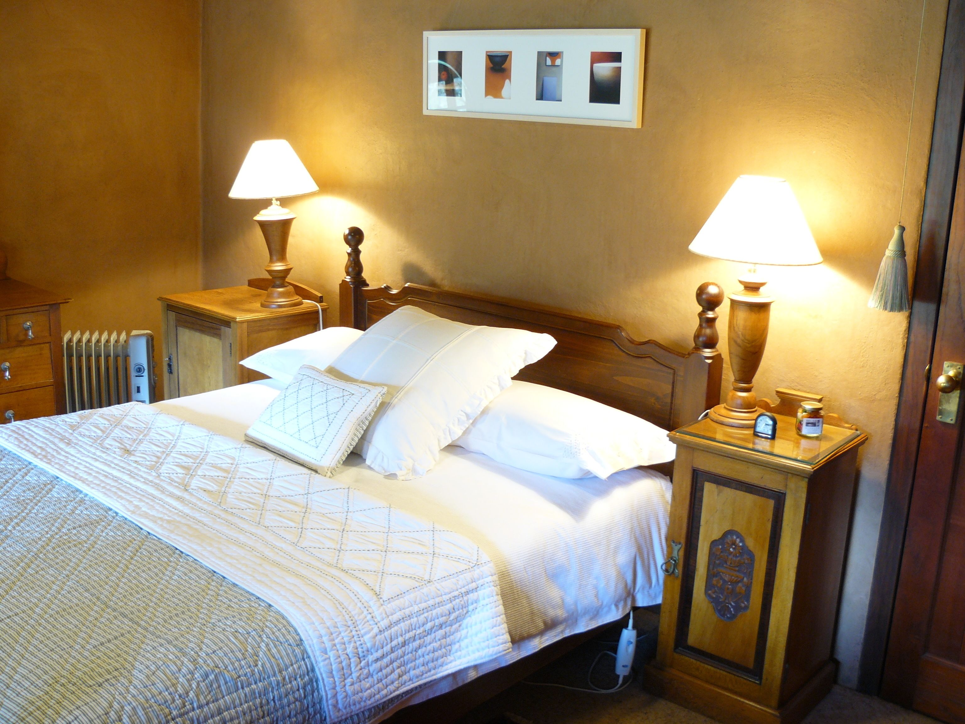 A neatly made bed with white linens and a quilt, flanked by two bedside tables with lamps, a radiator, and a framed picture on the wall.