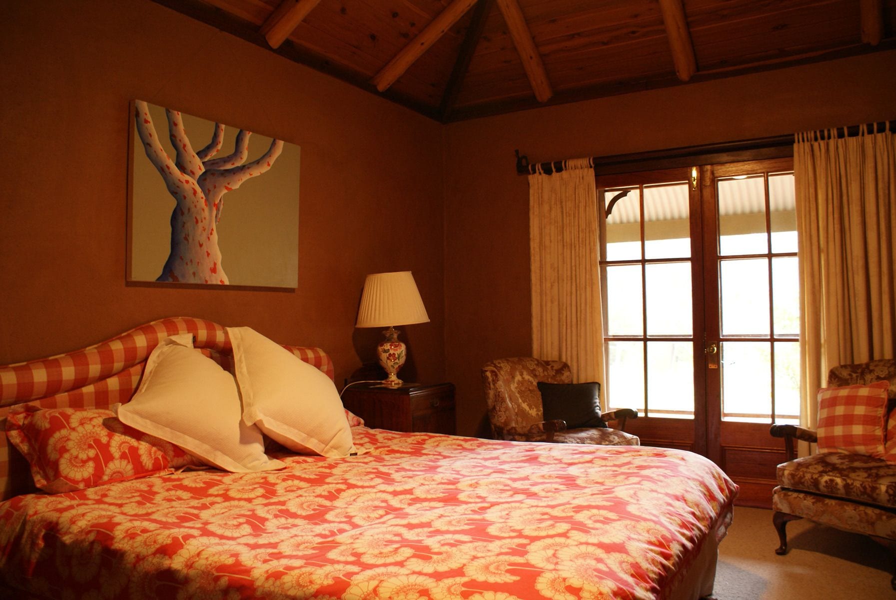 A bedroom with a large bed, a nightstand with a lamp, a chair, and a window with curtains.