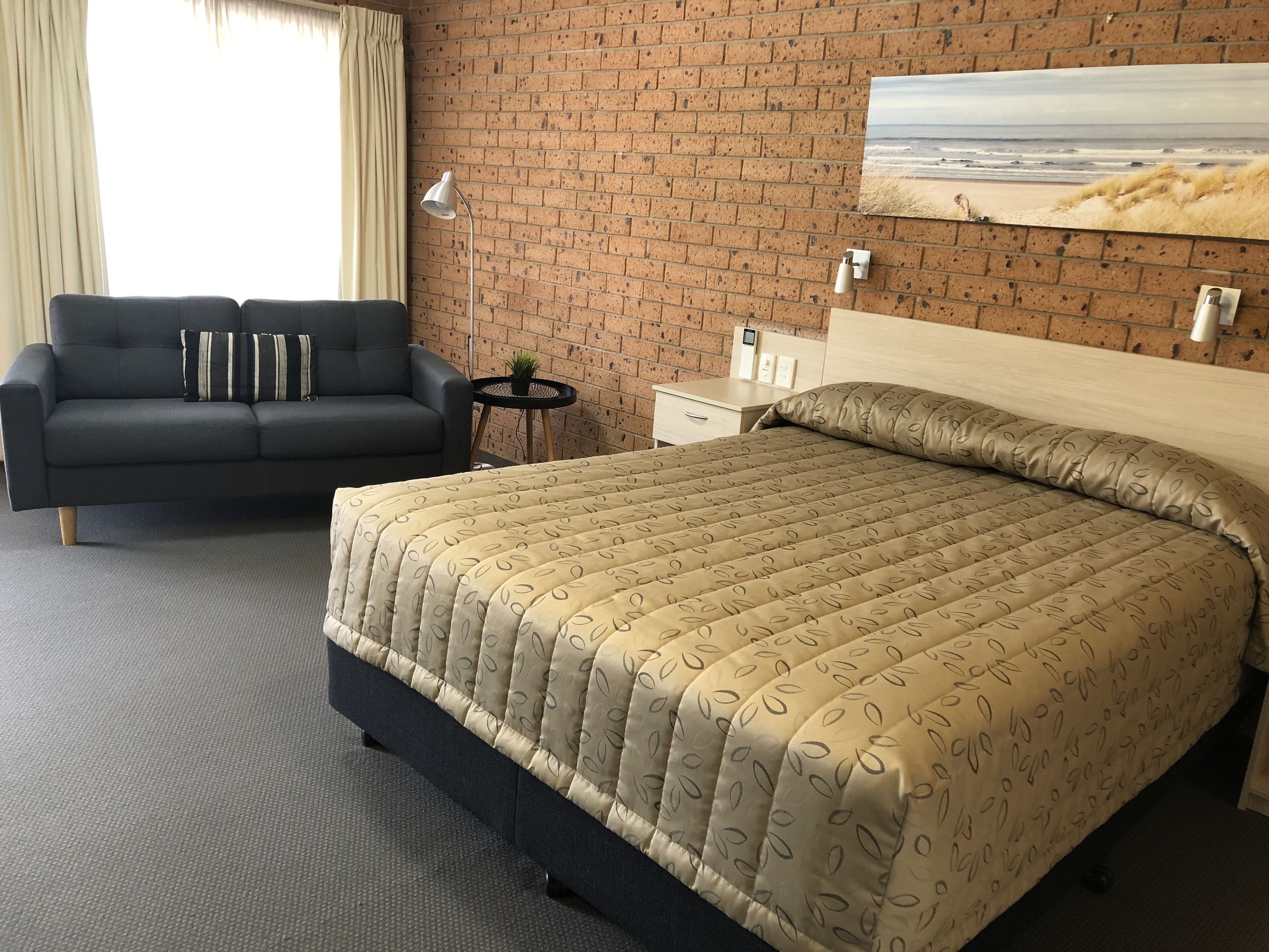 A hotel room with a brick wall, a bed with a patterned bedspread, a dark sofa with striped pillows, a small round table with a plant, and a painting of a beach scene.