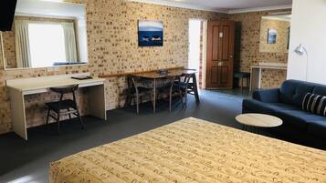 Quarto de hotel com cama, escrivaninha, cadeira, sofá, mesa pequena, televisão e porta.