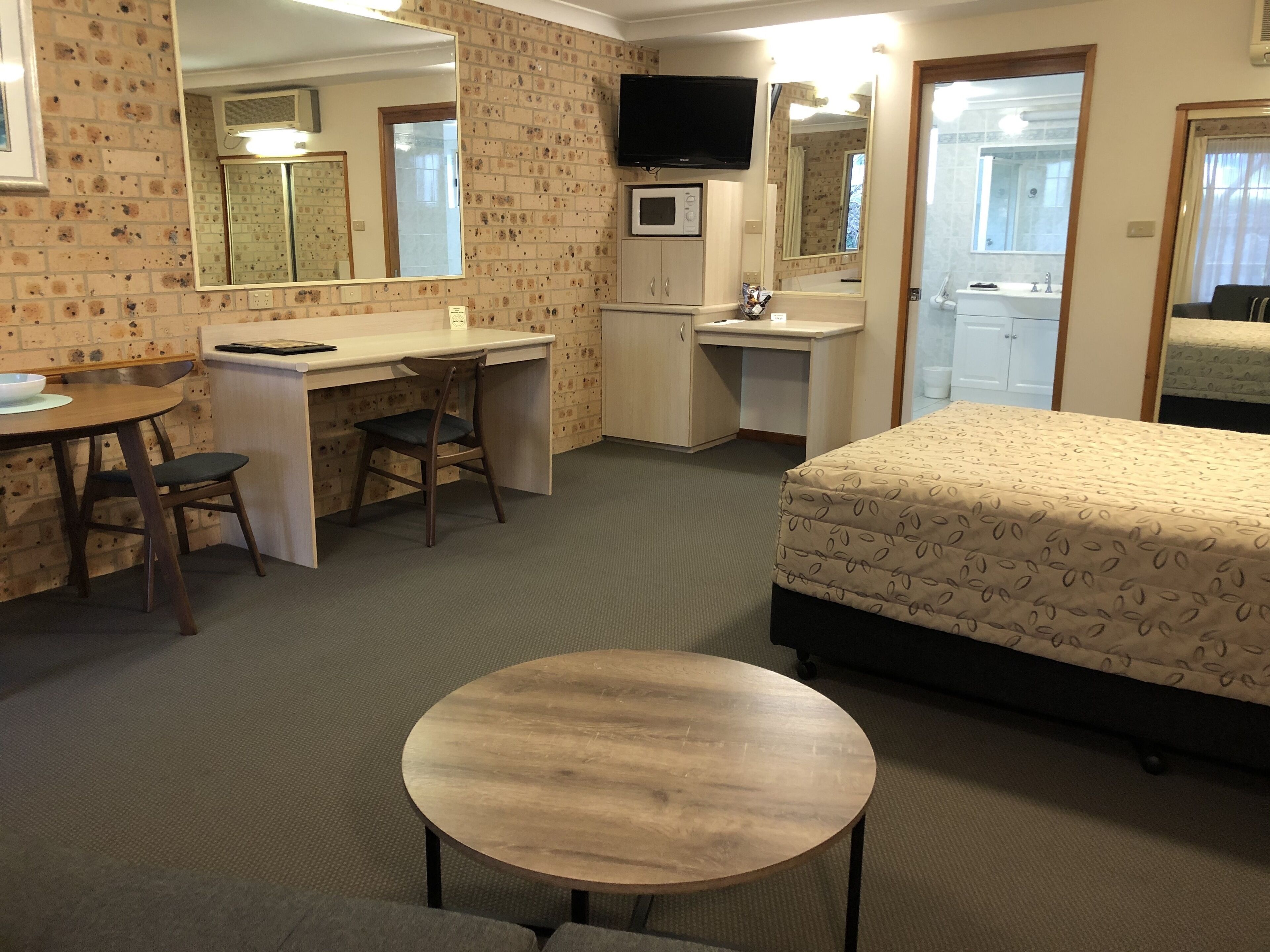 A compact hotel room with a bed, a desk, a chair, a small table, a TV, a microwave, and a bathroom visible through an open door.