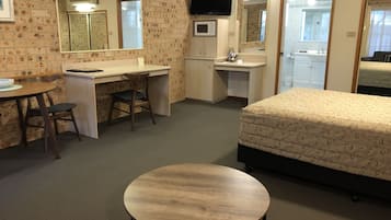 A compact hotel room with a bed, a desk, a chair, a small table, a TV, a microwave, and a bathroom visible through an open door.