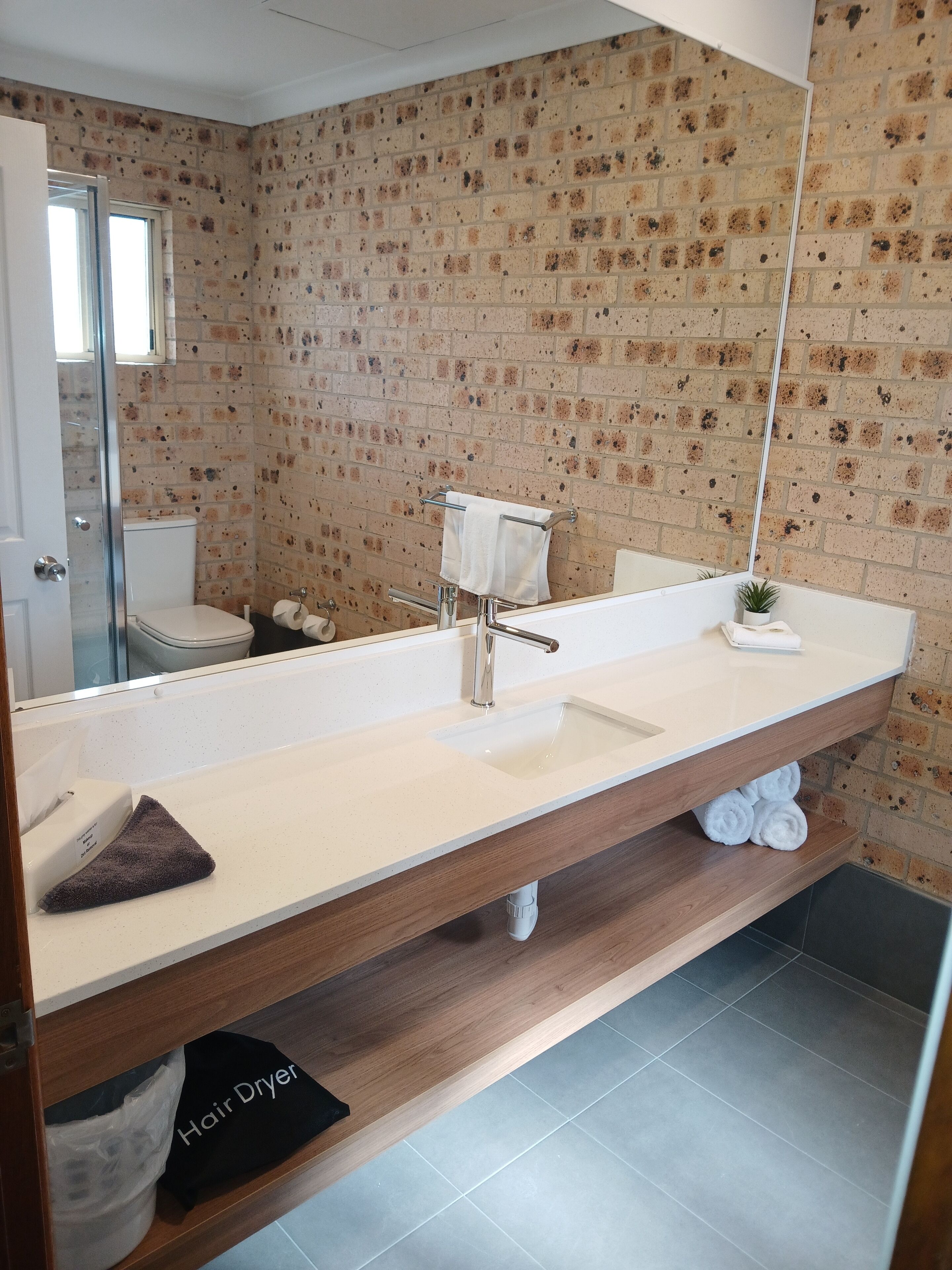 Un baño moderno con pared de ladrillo, un espejo grande, un lavabo blanco y un perchero para toallas.
