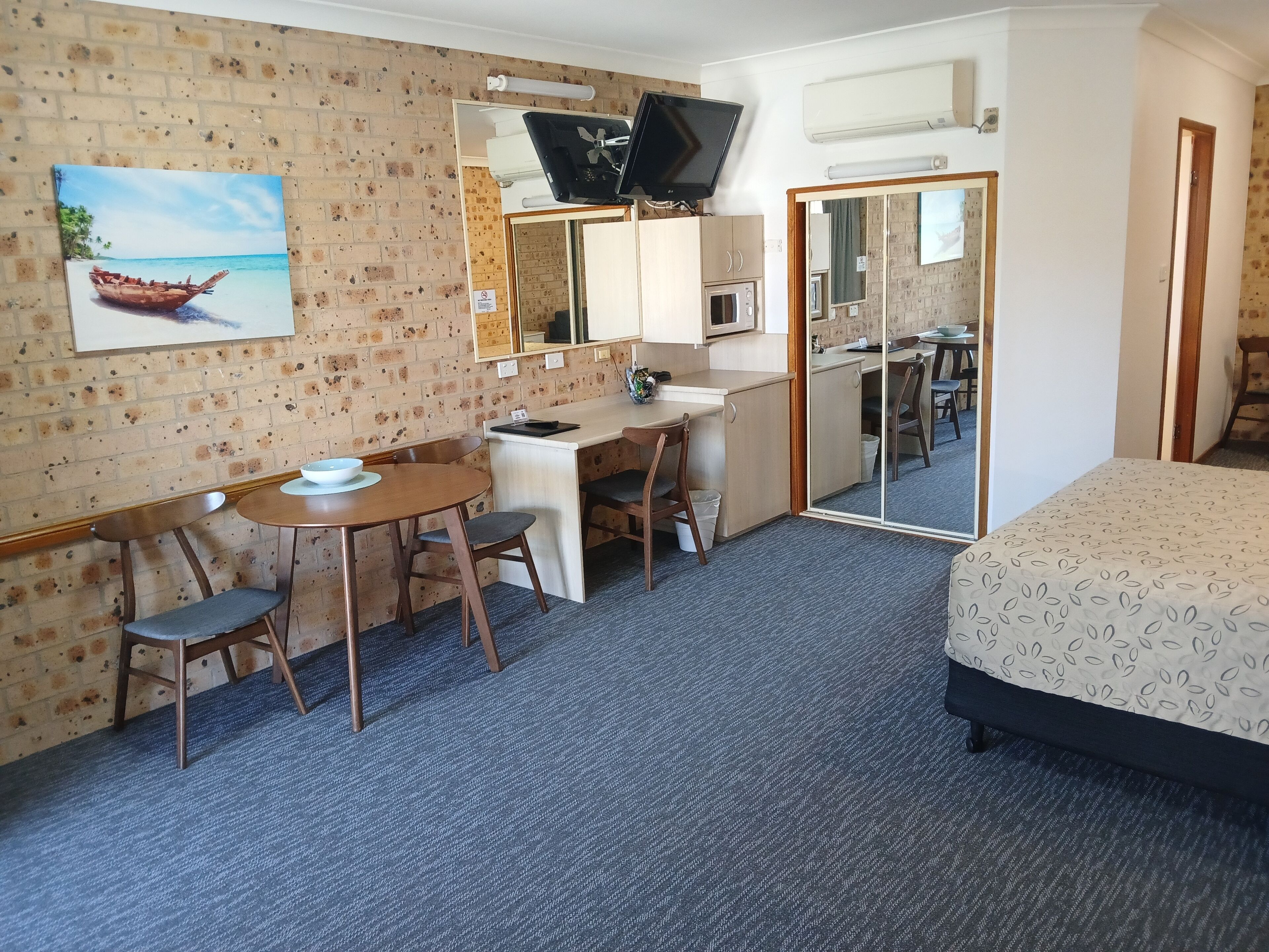 A hotel room with a bed, a desk with a chair, a small table with a bowl, a television, and a mirror.