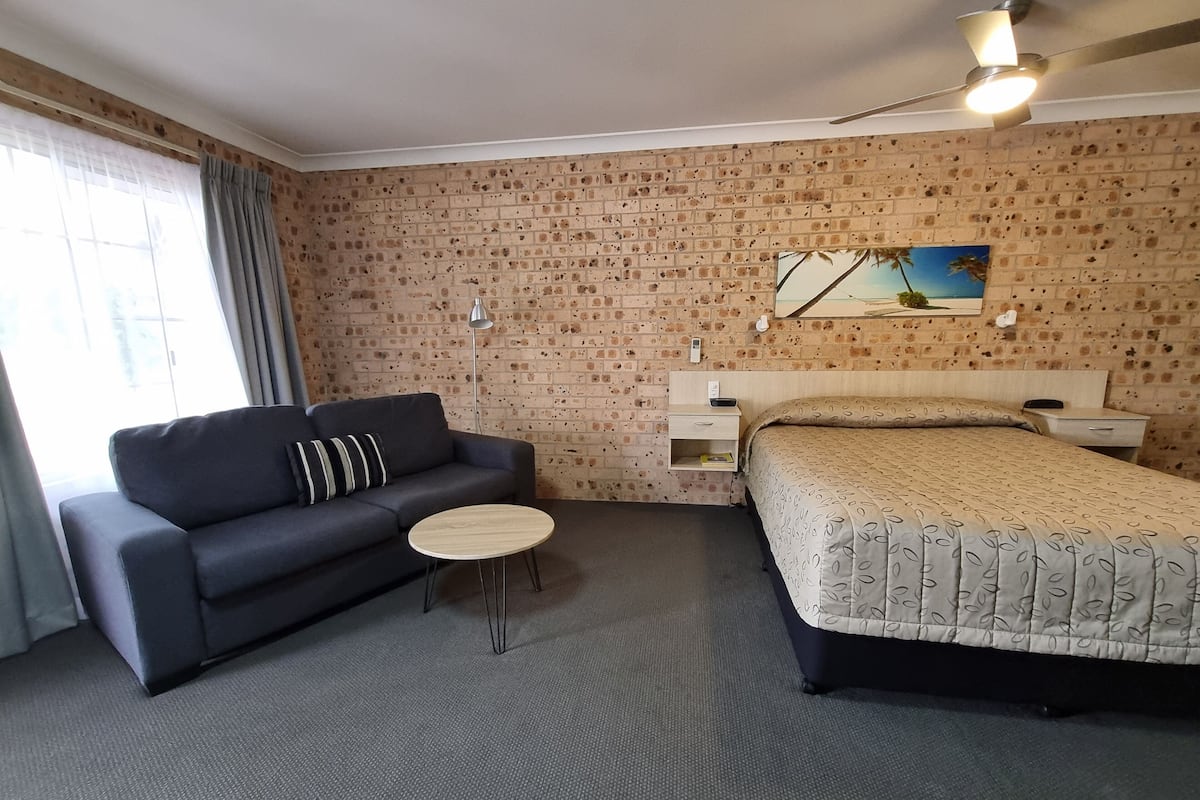 A hotel room with a brick-patterned wall, a bed with a patterned bedspread, a dark sofa with a striped pillow, a small round table, and a nightstand with a lamp.
