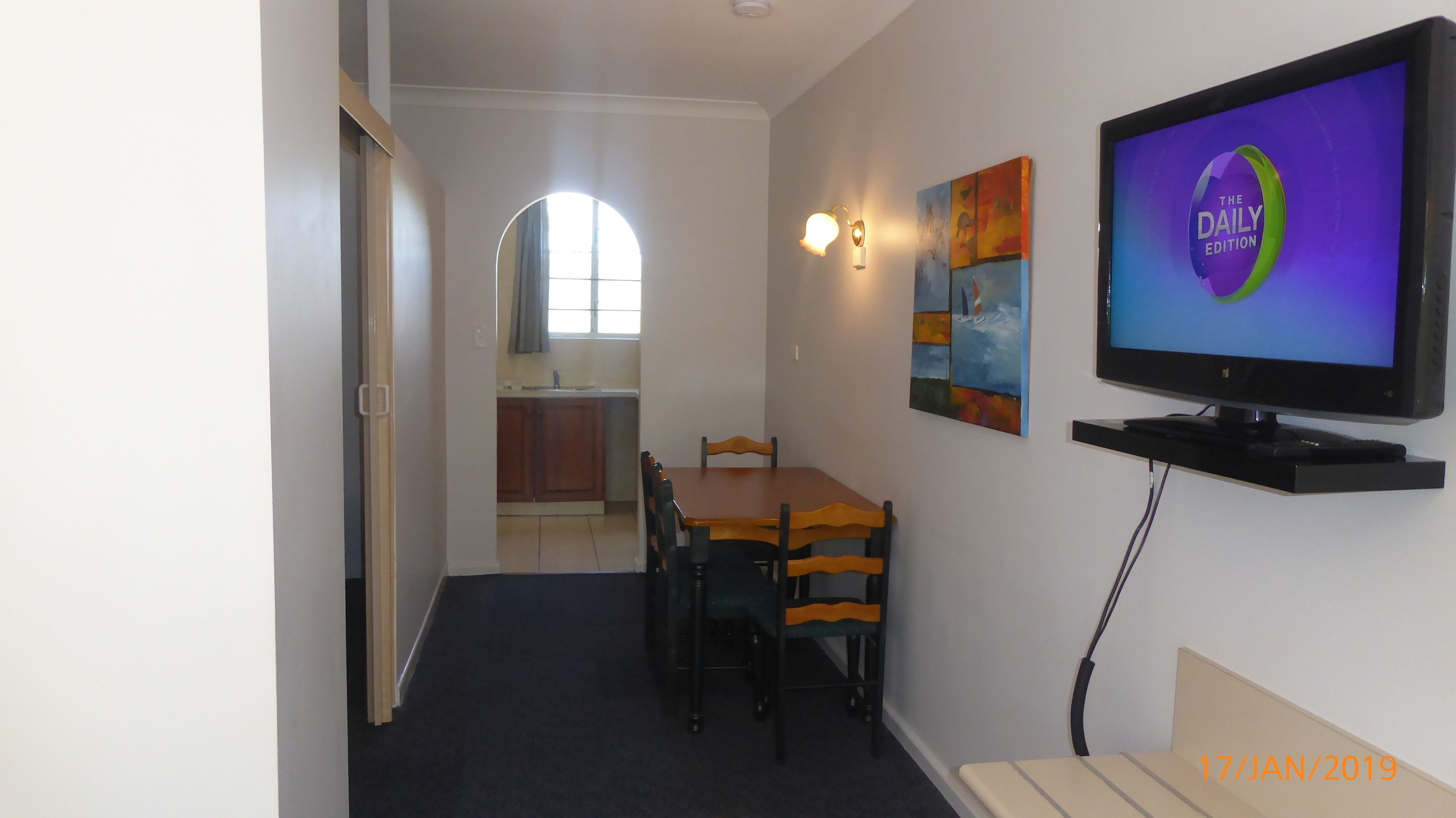 A compact living space with a dining area, a television mounted on the wall, and a kitchen in the background.