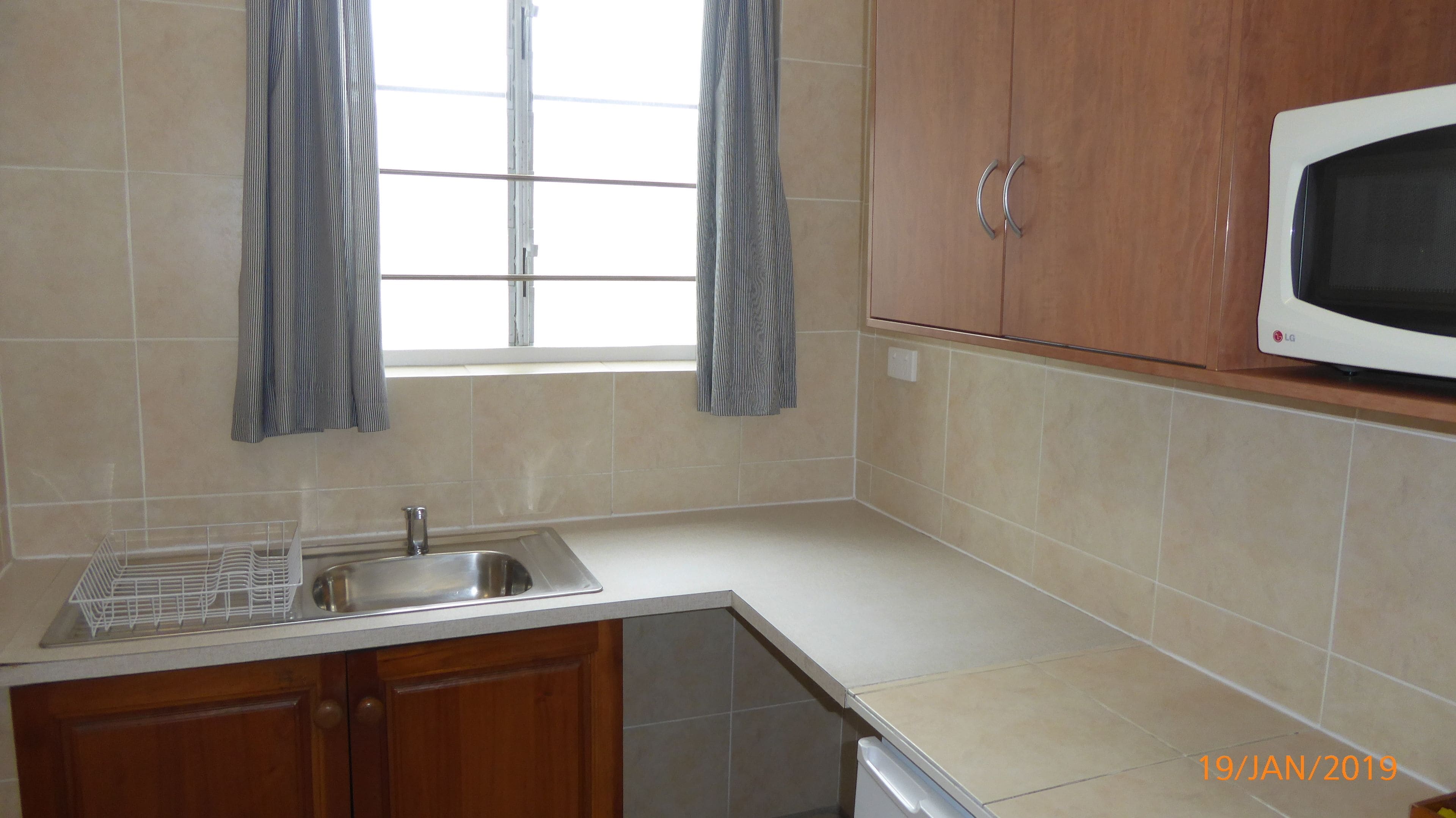 Una cocina con un lavamanos de acero inoxidable, armarios de madera, un microondas y una ventana con cortinas.