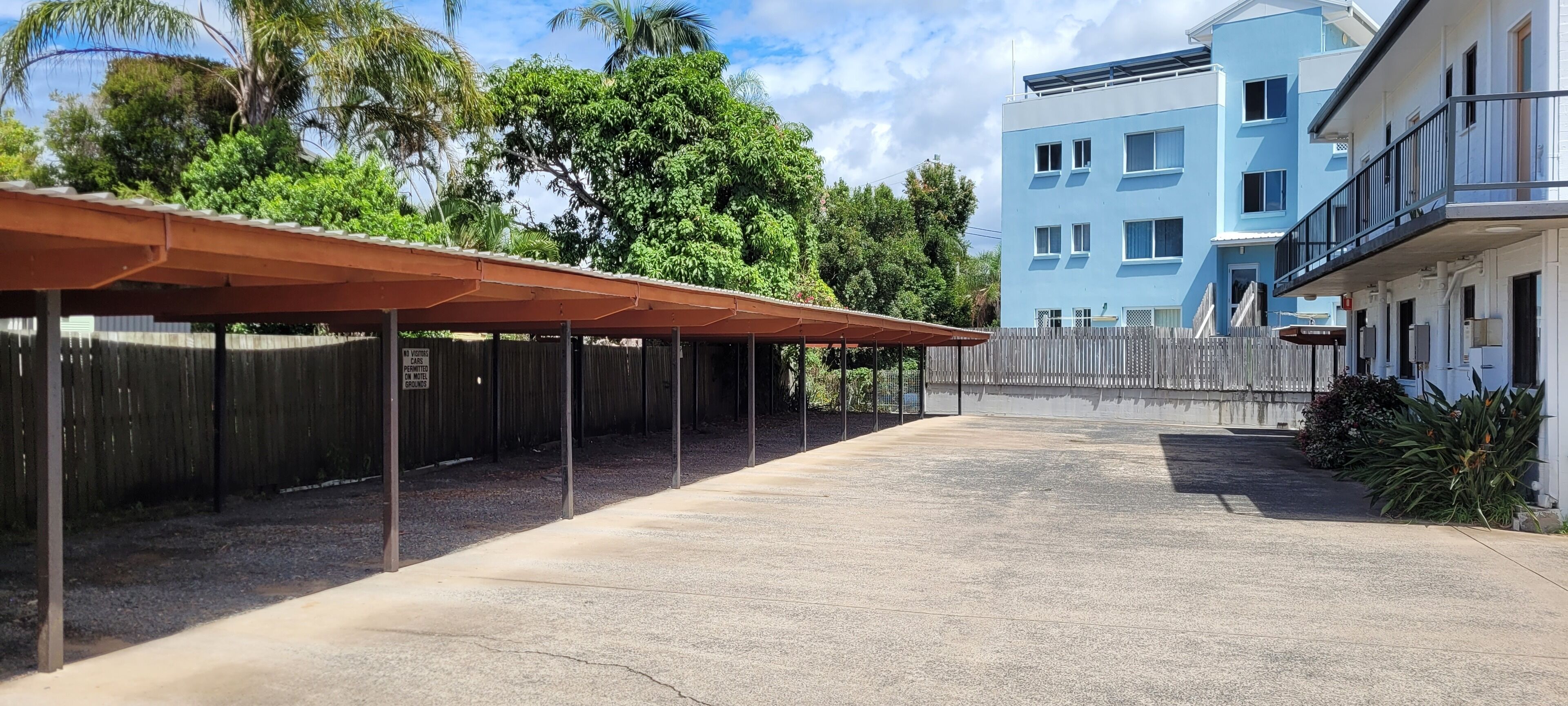 A covered outdoor area with a concrete floor, surrounded by a wooden fence and a building with balconies.