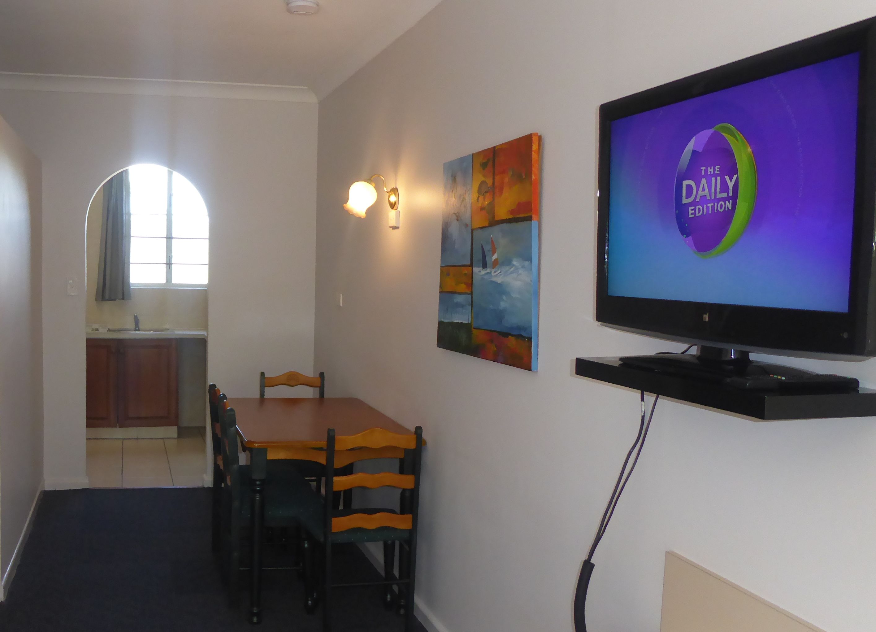 A dining area with a wooden table and chairs, a television displaying 'The Daily Edition', and a kitchen in the background.