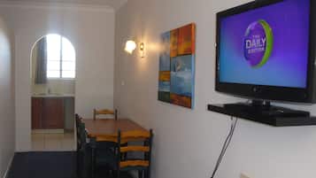 A dining area with a wooden table and chairs, a television displaying 'The Daily Edition', and a kitchen in the background.