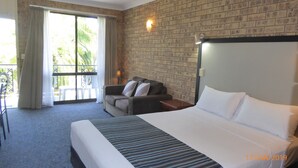 A hotel room with a large bed, a sofa with pillows, a small table, and a balcony with a view of greenery. - Tower Court Motel (Torquay)