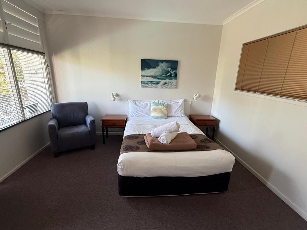 Una habitación de hotel con una cama, una silla, una mesita de noche y una ventana con persianas.