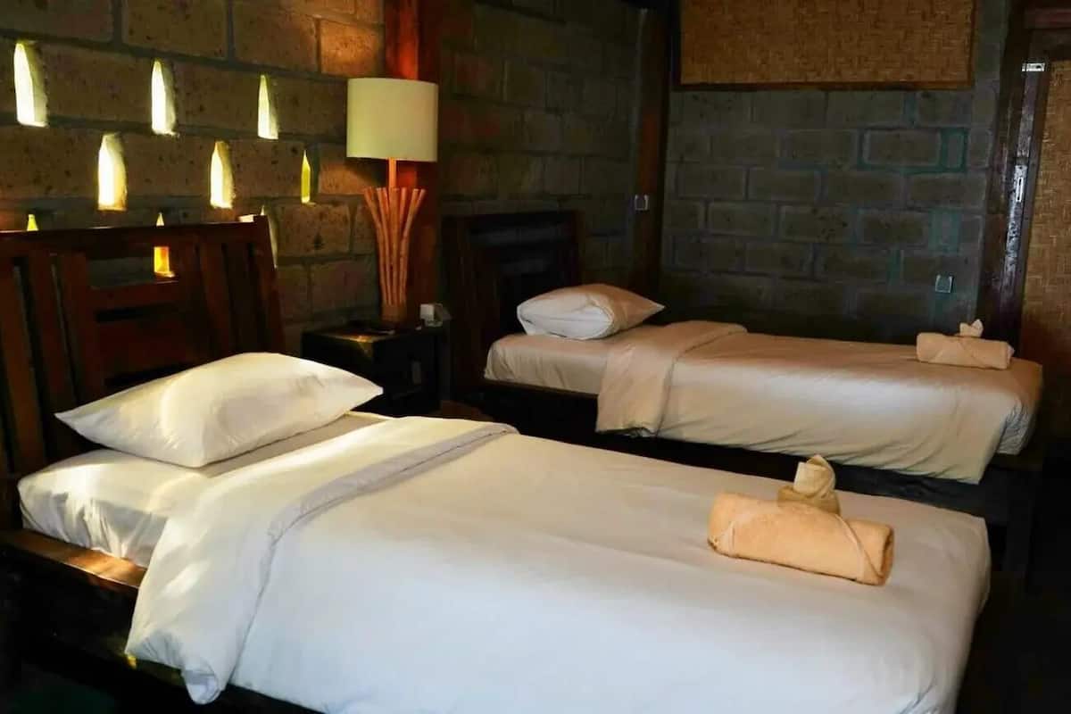 A hotel room with two beds, a wooden headboard, a nightstand, a lamp, and a brick wall.