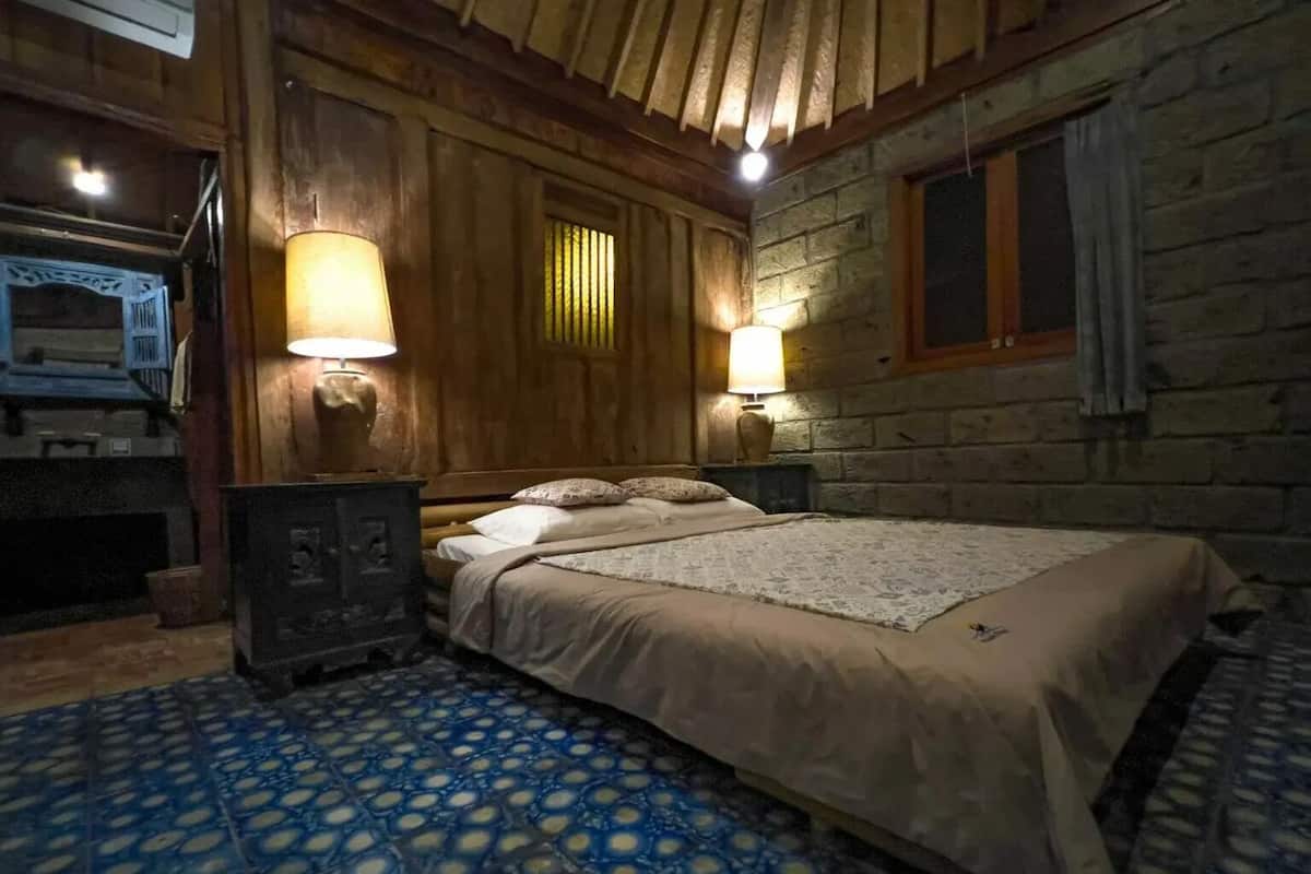 A cozy room with a bed, two wall-mounted lamps, a wooden headboard, and a stone wall.