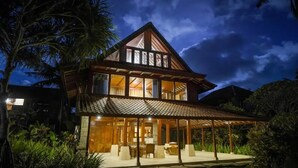 Uma casa de dois andares com varanda coberta, cercada por vegetação tropical.
