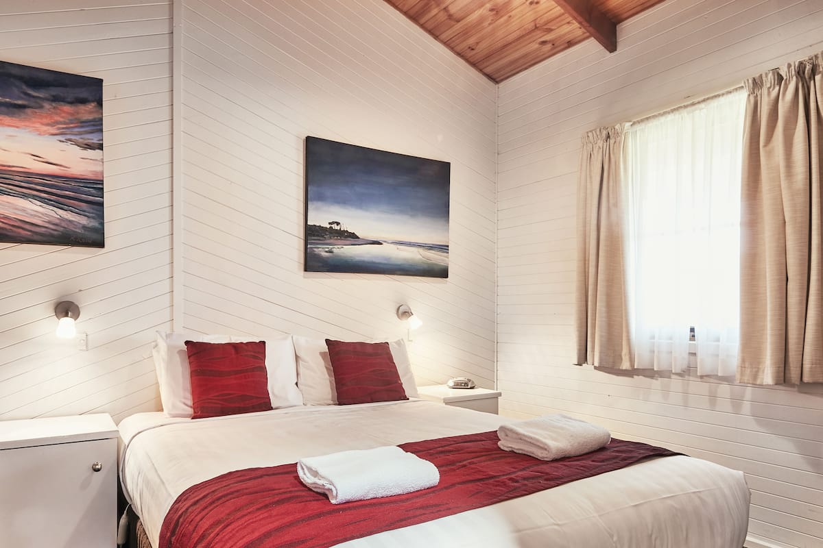 Un dormitorio ordenado con cama, mesita de noche, un cuadro en la pared y ventana con cortinas.