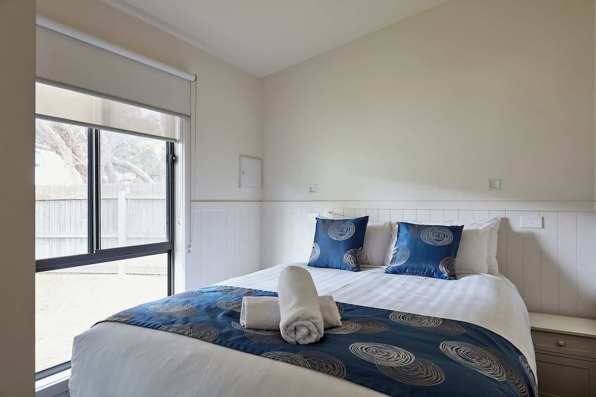 Habitación de hotel con una cama grande, una ventana con persianas, una mesita de noche y un interruptor de luz en la pared.