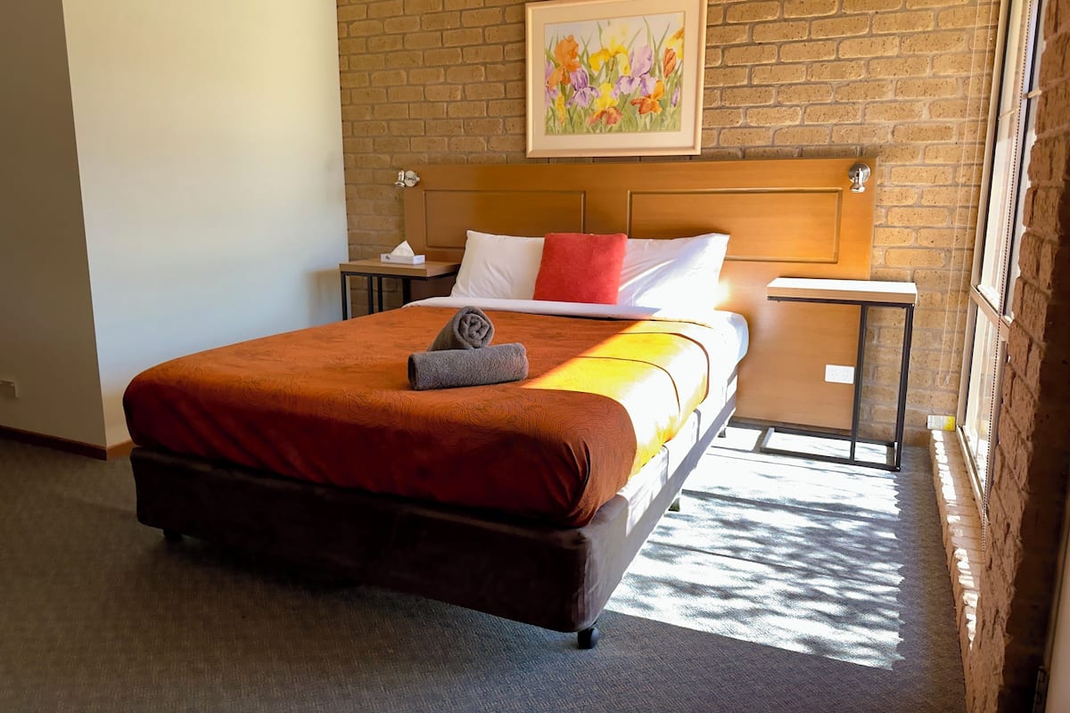 A hotel room with a brick wall, a bed with a brown headboard, white and red pillows, a rolled towel, a nightstand with a tissue box, and a framed painting above the bed.