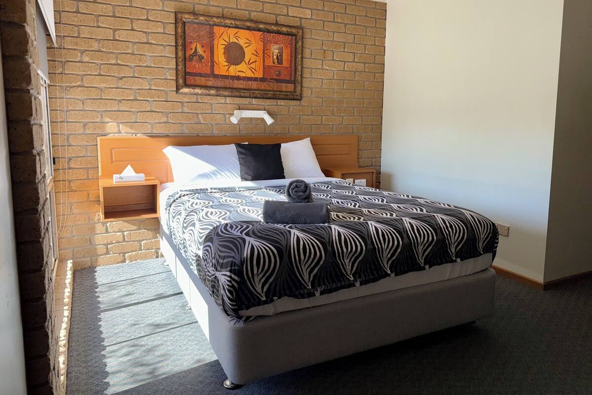 A hotel room with a brick wall, a bed with patterned bedding, a nightstand, and a wall-mounted artwork.
