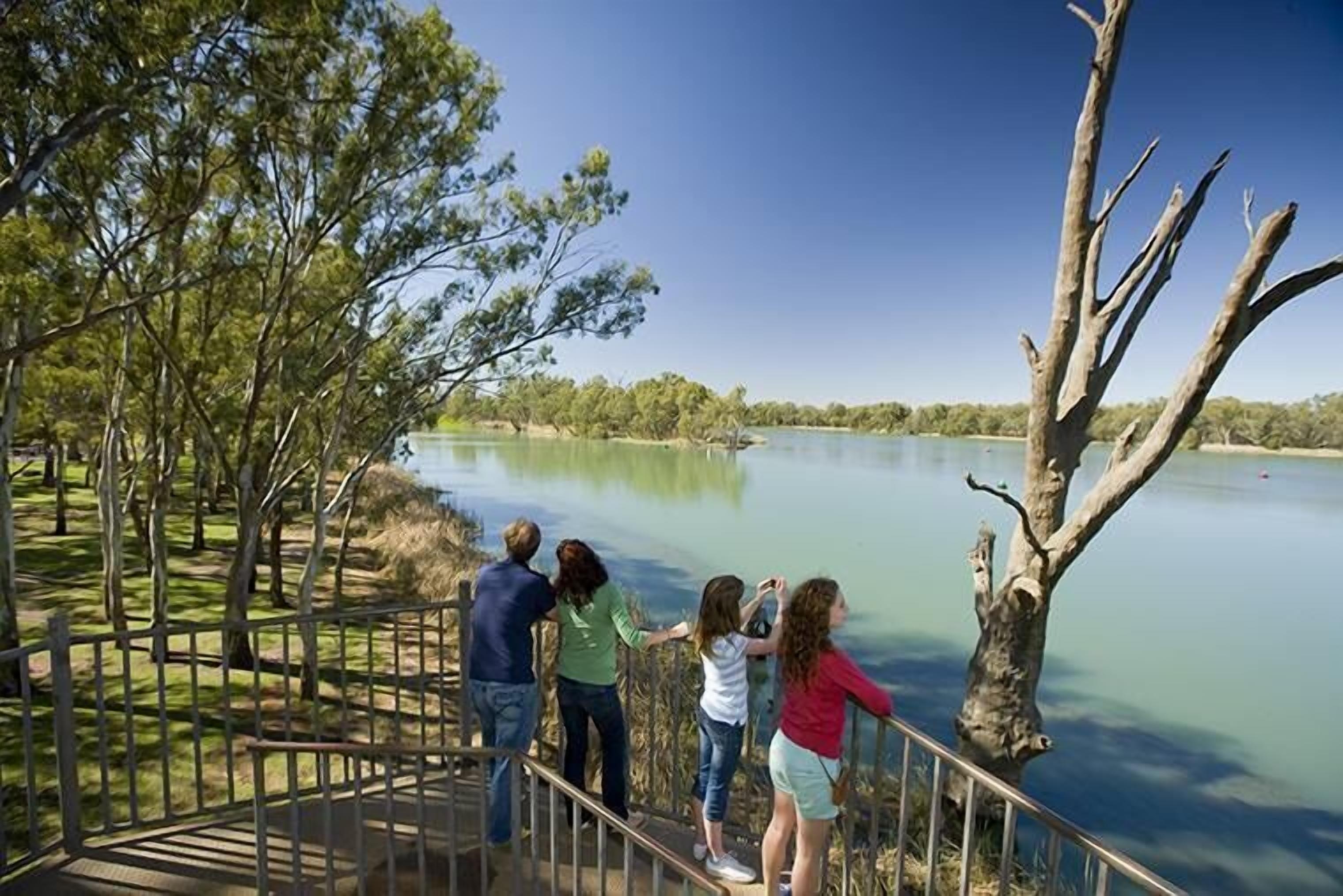 Un grupo de personas está de pie en una cubierta de madera, con vista a un lago tranquilo, con árboles y un cielo despejado al fondo.
