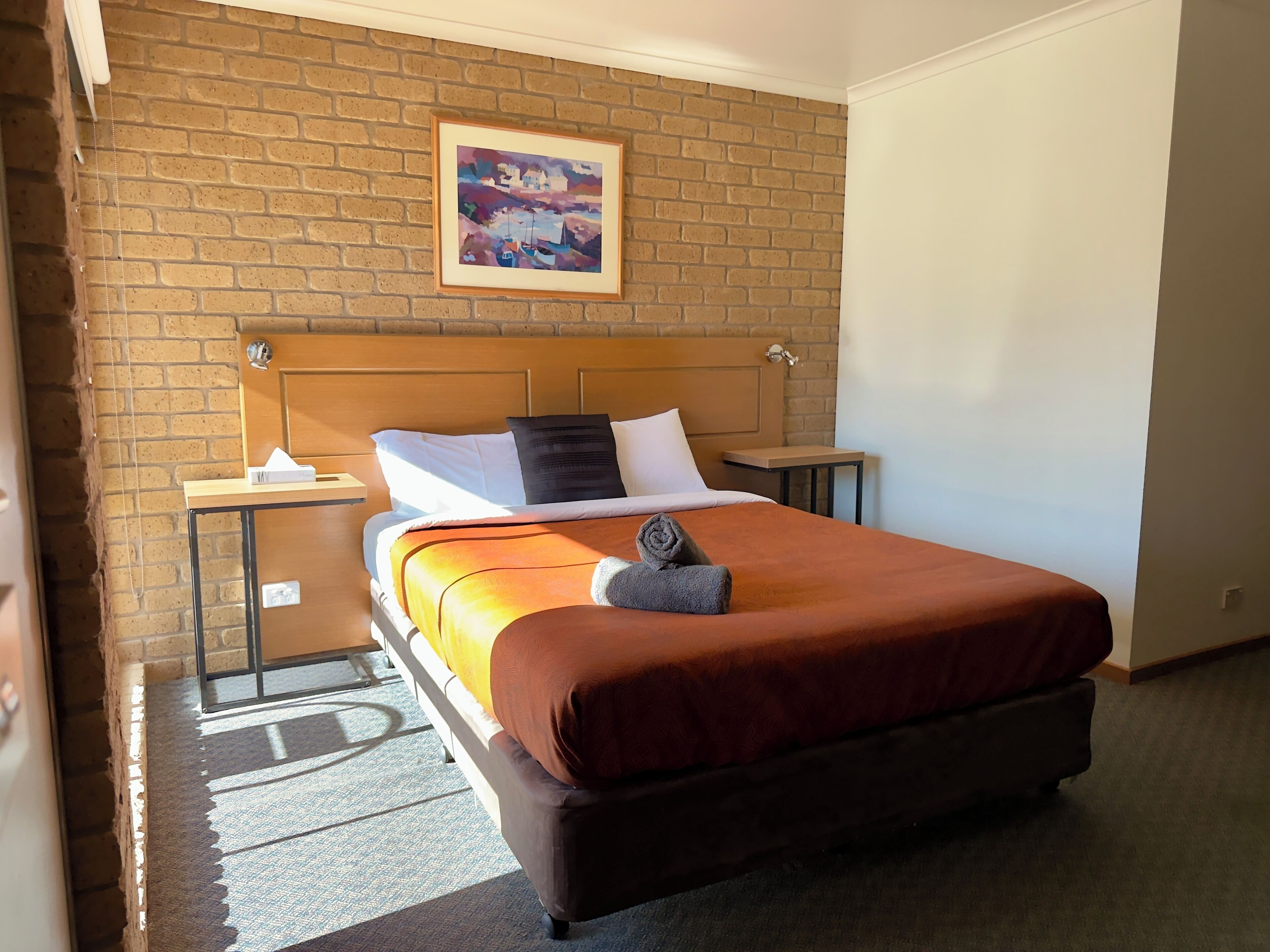A hotel room with a brick wall, a bed with a wooden headboard, a nightstand, a table, and a painting above the bed.