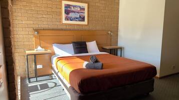 A hotel room with a brick wall, a bed with a wooden headboard, a nightstand, a table, and a painting above the bed.