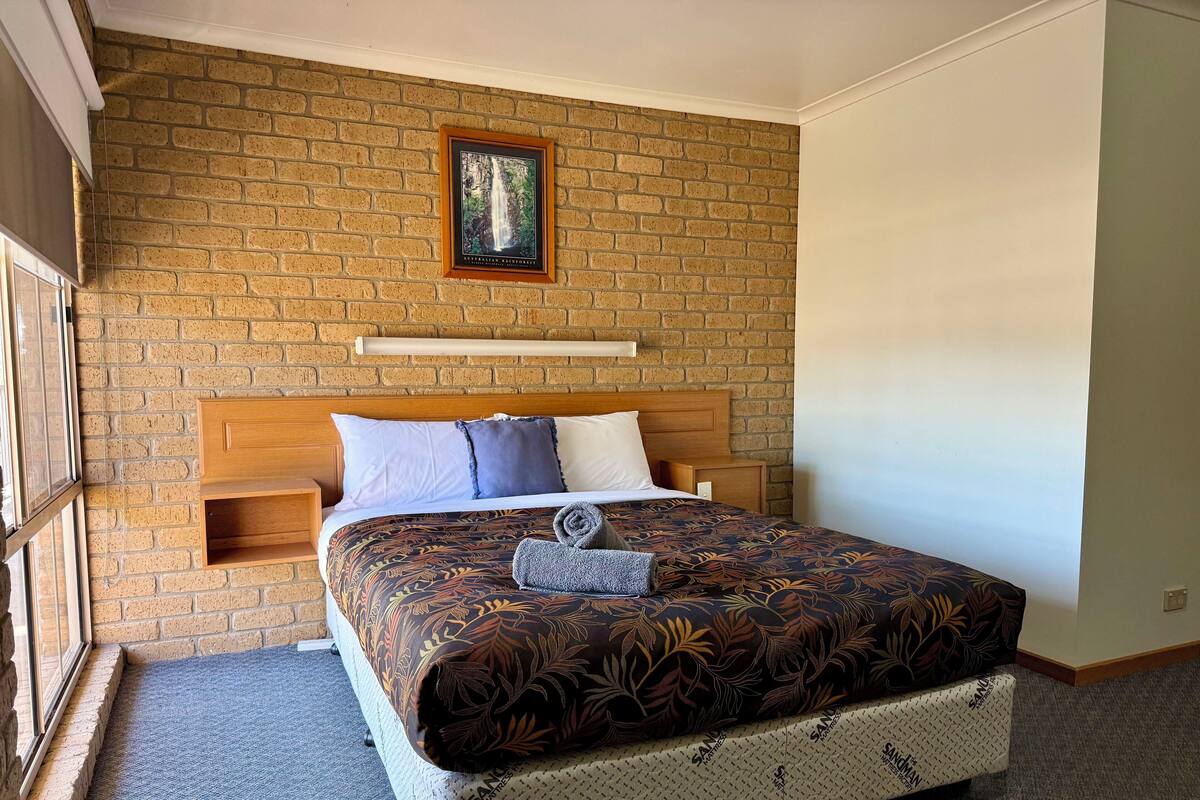 A hotel room with a brick wall, a framed picture of a waterfall, a bed with patterned bedding, and a small wooden shelf.